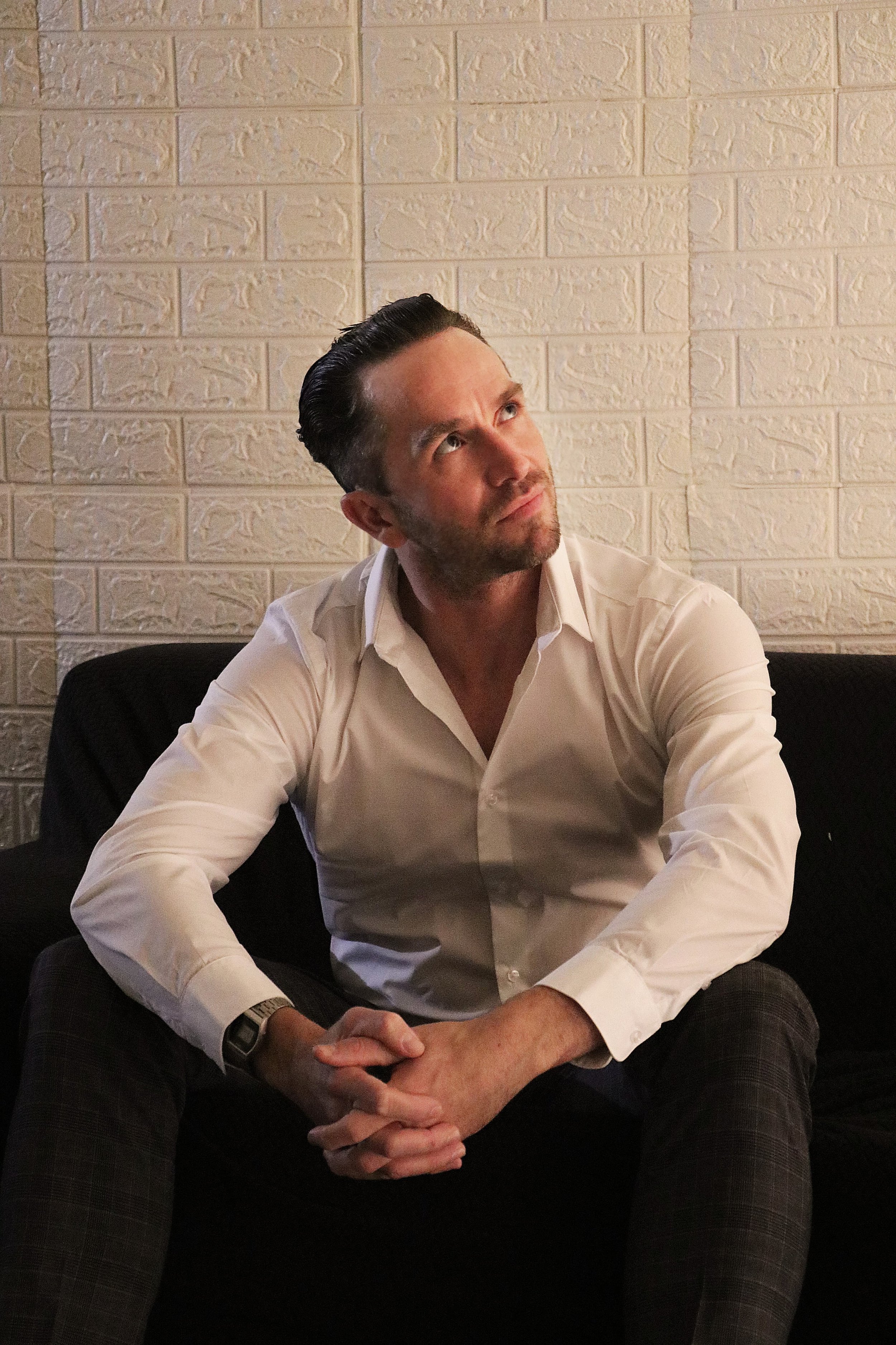 A man with dark, slicked-back hair, wearing a white dress shirt, sitting in front of a cream-colored brick wall, looking upwards and to his left, with his hands clasped. The man appears to be indoors.