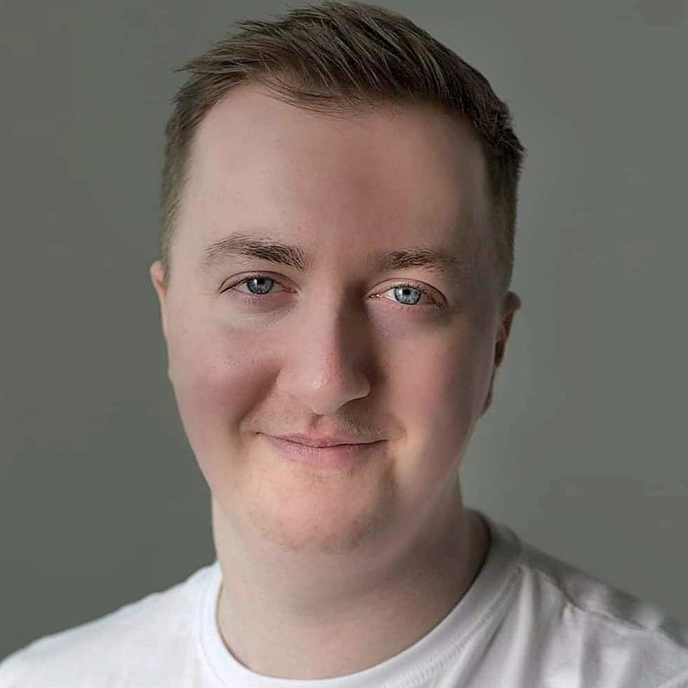 Close-up of a smiling young man with short brown hair and blue eyes, wearing a white shirt against a plain wall.