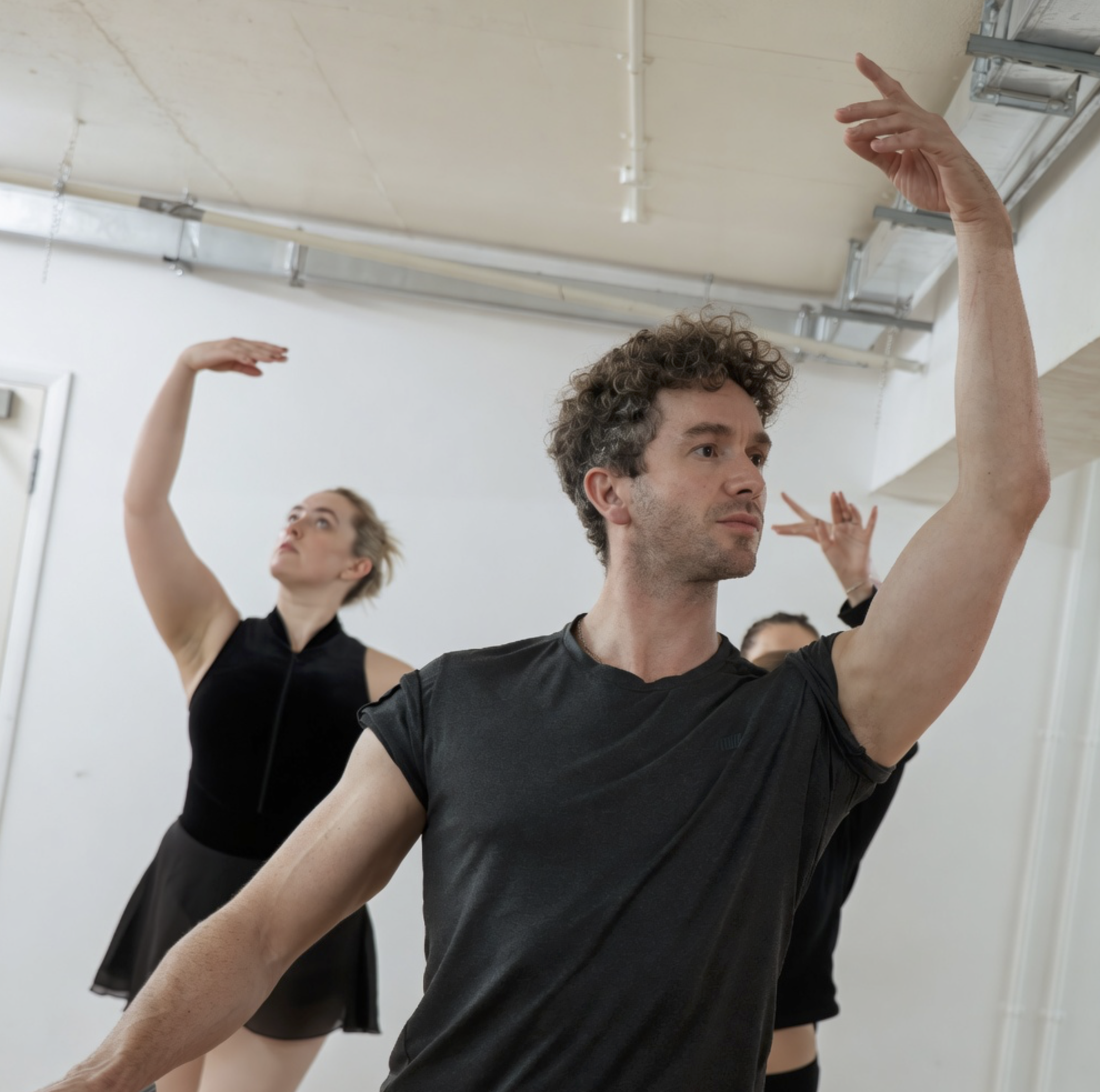 People practicing ballet, raising their right arms above their heads in a dance studio.