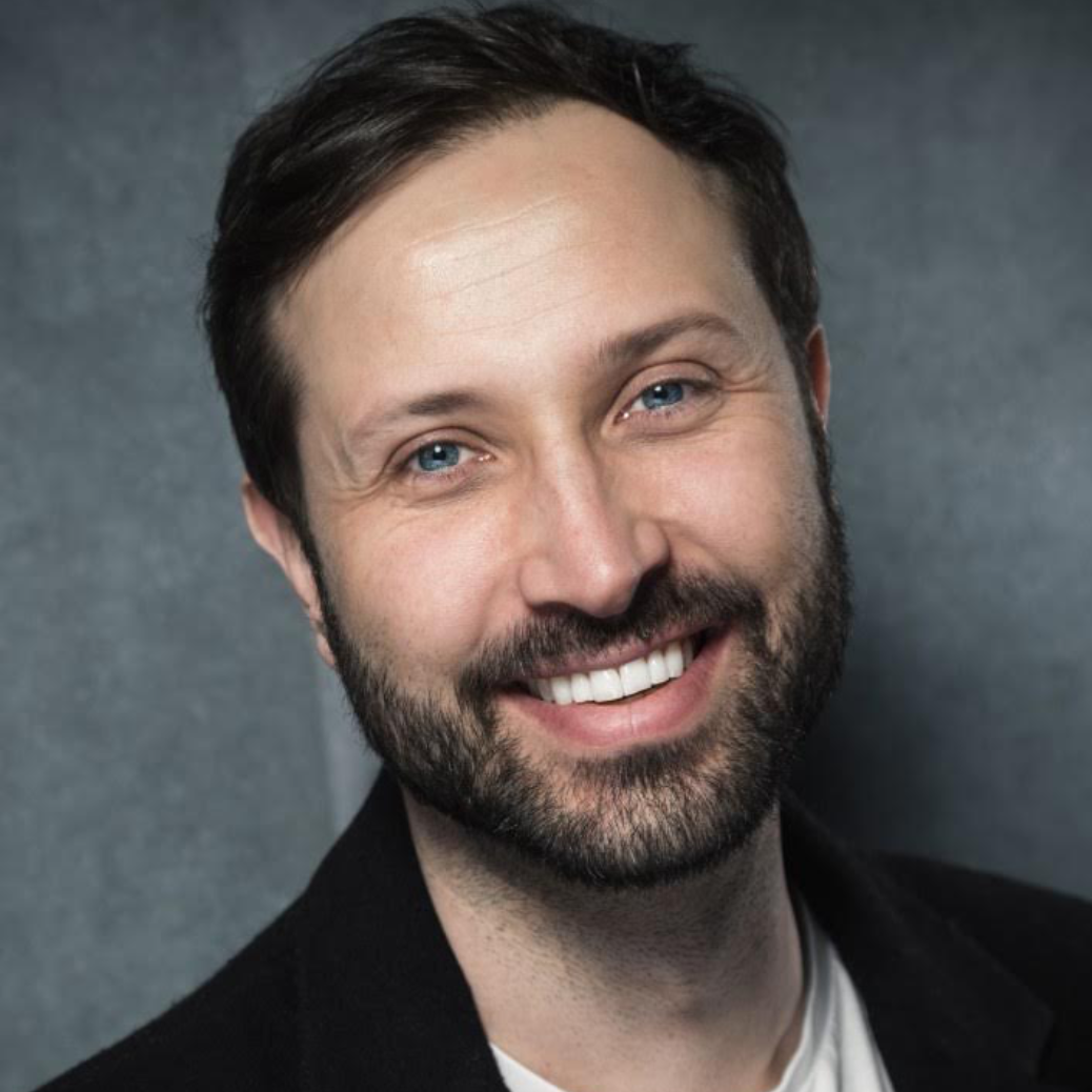 Portrait of a smiling man with short dark hair, blue eyes, and a beard, wearing a black blazer and white shirt, against a gray background.