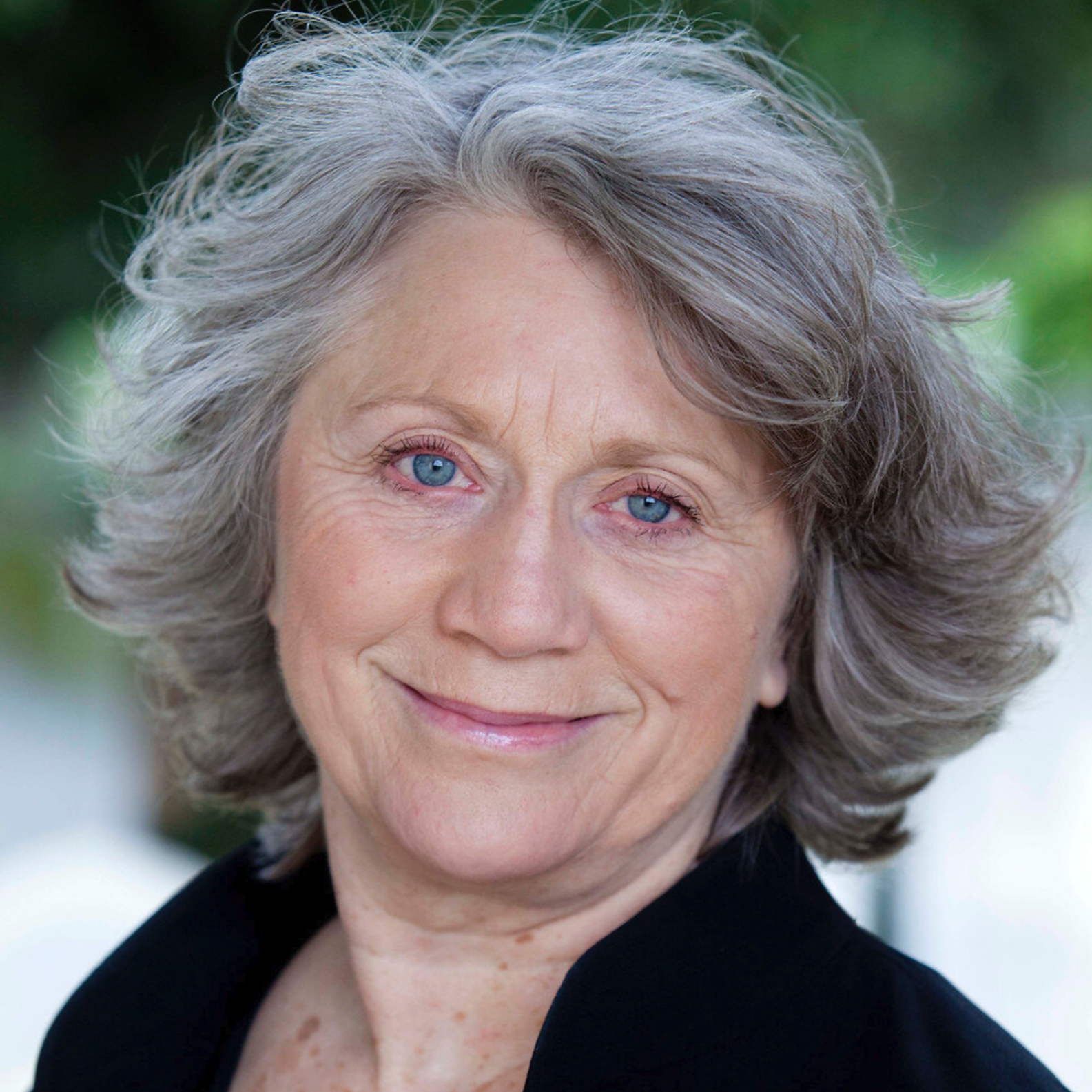 Close-up of an elderly woman with gray, curly hair, blue eyes, and a gentle smile, wearing a black top against a blurred outdoor background.