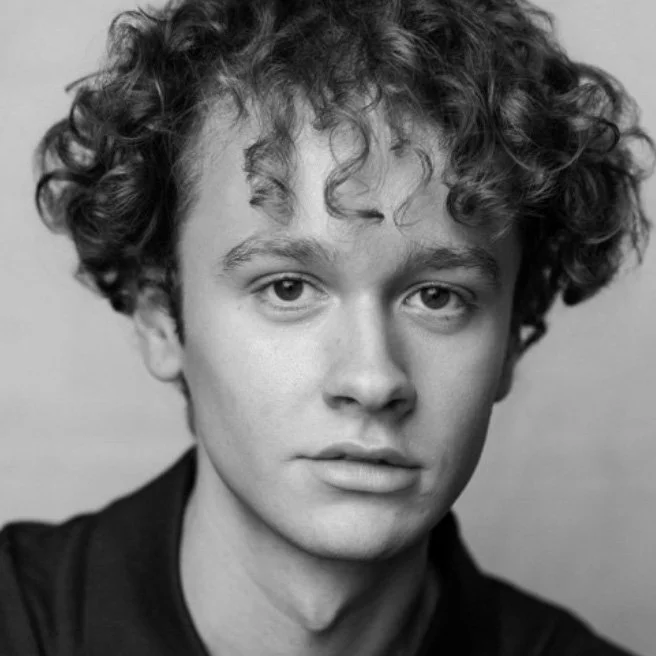 Black and white close-up portrait of a young person with curly hair and light skin, wearing a dark top.