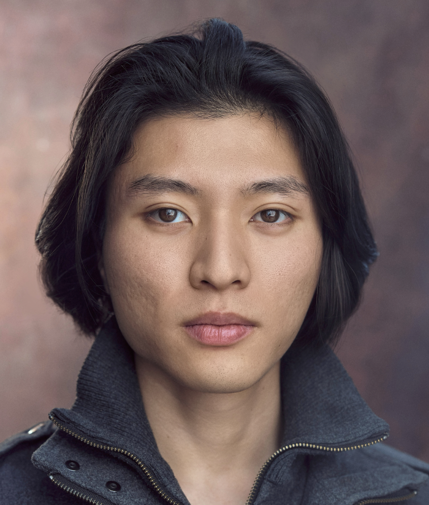 Close-up portrait of a young woman with short black hair, wearing a dark zip-up jacket, against a blurred, warm-toned background.