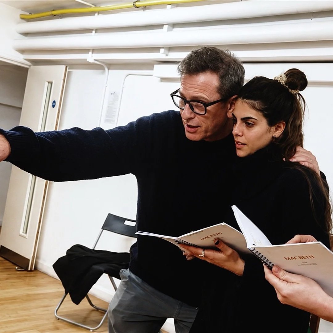 Two people, a man and a woman, are close together looking and pointing at the same direction. The woman is holding a script of Macbeth, and they seem to be rehearsing or discussing a scene.