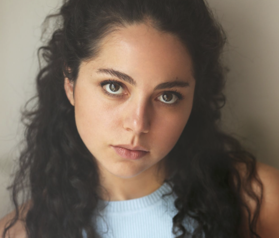 Close-up portrait of a young woman with dark curly hair and fair skin, wearing a light blue top, looking directly at the camera with a neutral expression.