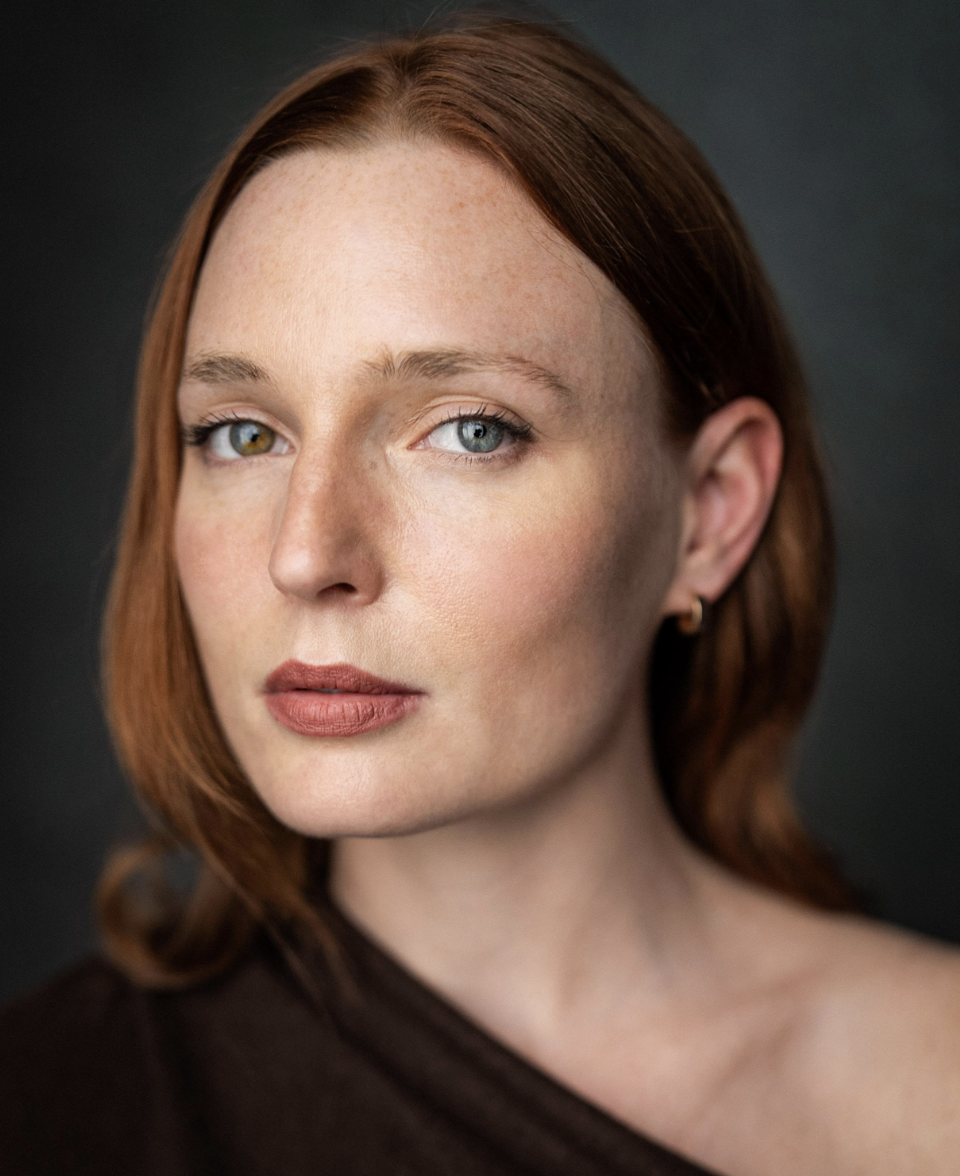Close-up portrait of a woman with red hair, fair skin with freckles, blue eyes, wearing minimal makeup, and a dark top, against a dark background.