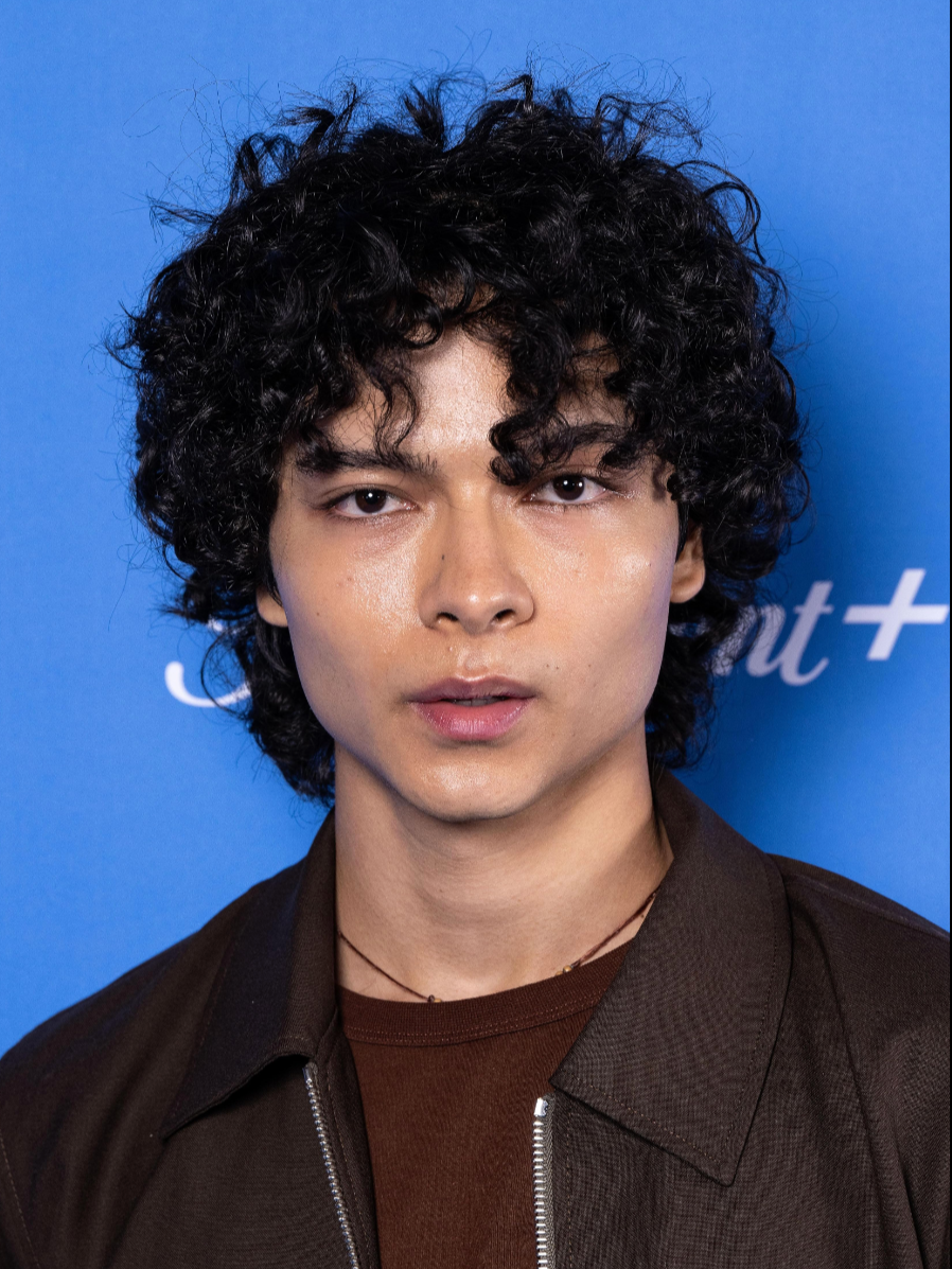 Close-up of a young person with curly black hair, wearing a brown shirt and black jacket, against a blue background with partially visible white text.