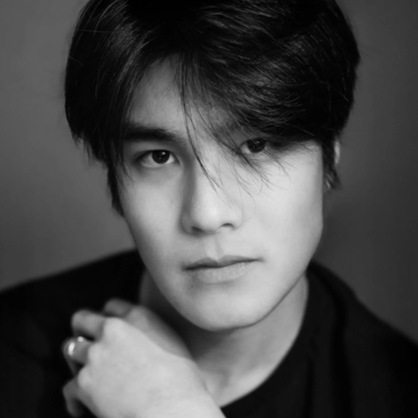Black and white close-up portrait of a young man with dark hair, partially covering one eye, wearing a dark top, with his hand resting on his shoulder.