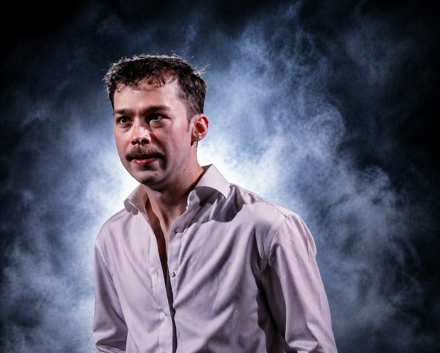 A man with short dark hair, a mustache, and fair skin, wearing a light-colored dress shirt, standing against a dark background with smoke or fog behind him.