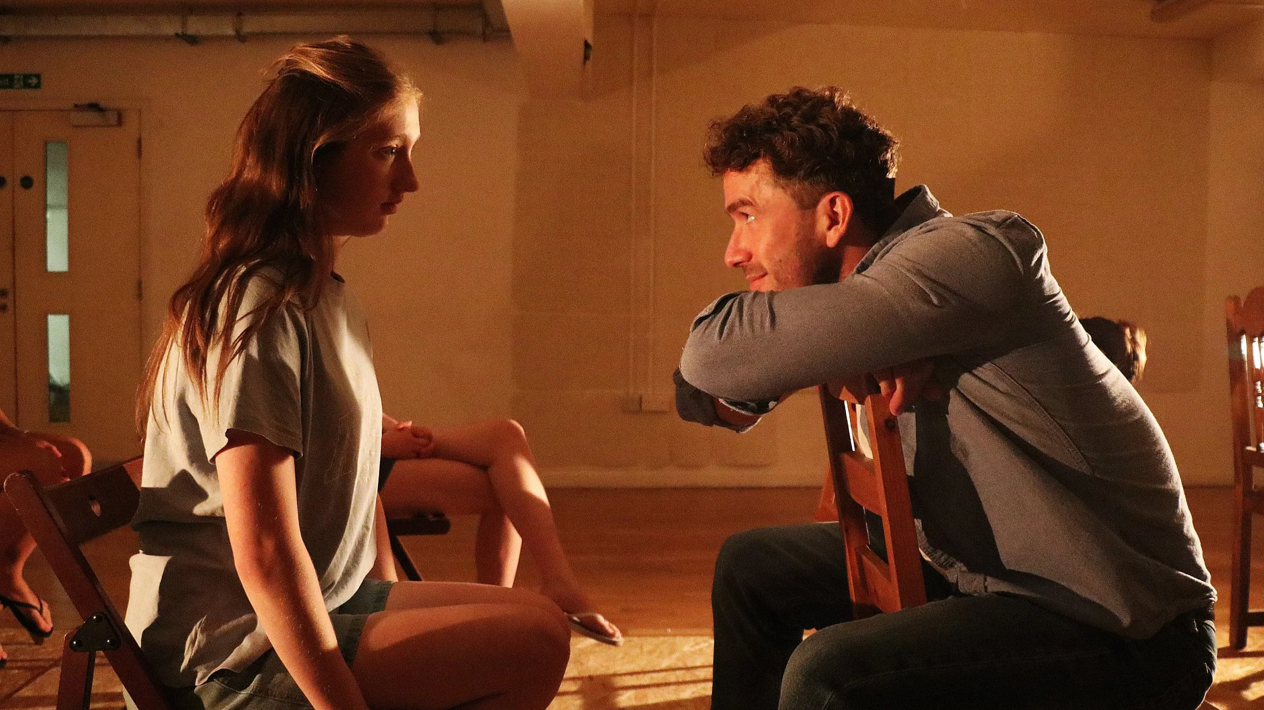 A young woman sitting on a wooden chair, facing a man kneeling in front of her. The man has his arms resting on the back of a chair, and they appear to be having an intense conversation in a warmly lit room with wooden flooring.