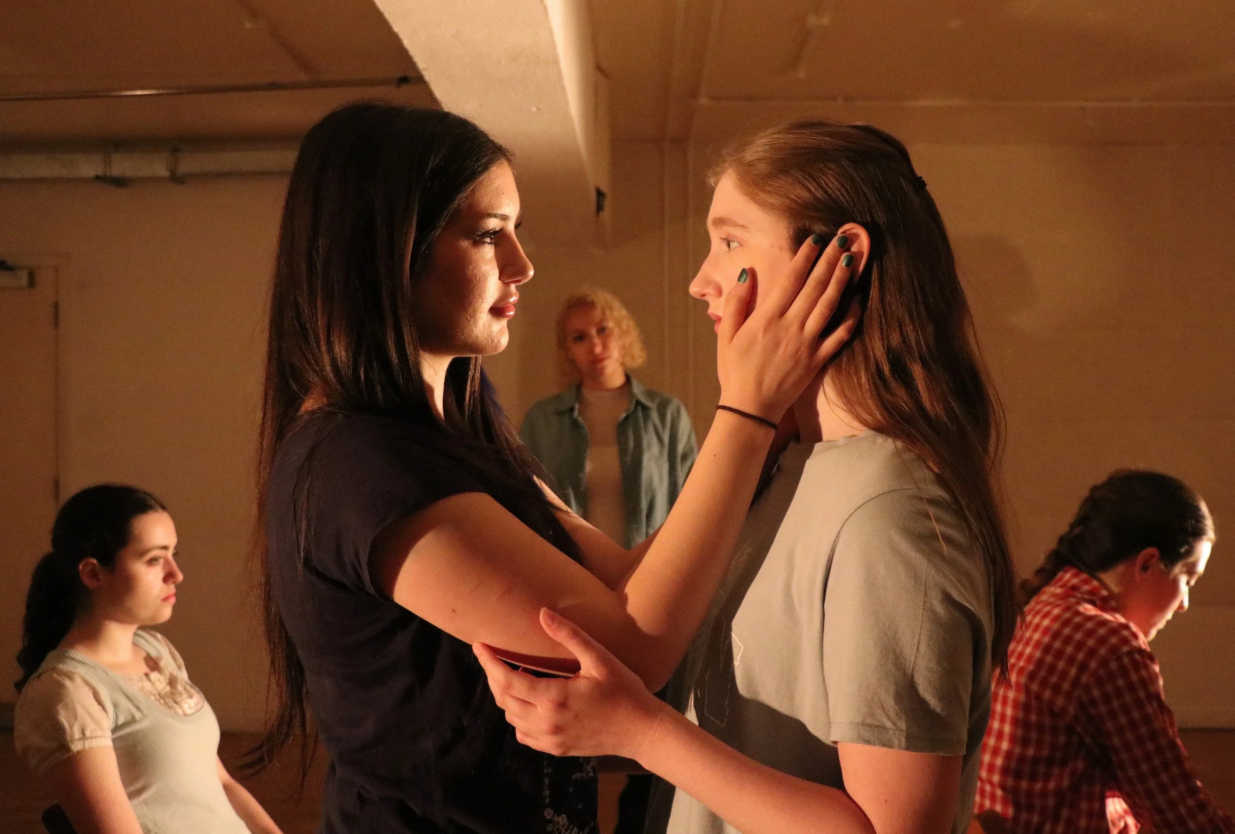 Two young women are standing close to each other, gazing into each other's eyes. The woman on the left has long dark hair and is touching the face of the woman on the right, who has long brown hair. In the background, there are several people, mostly women, sitting and observing.