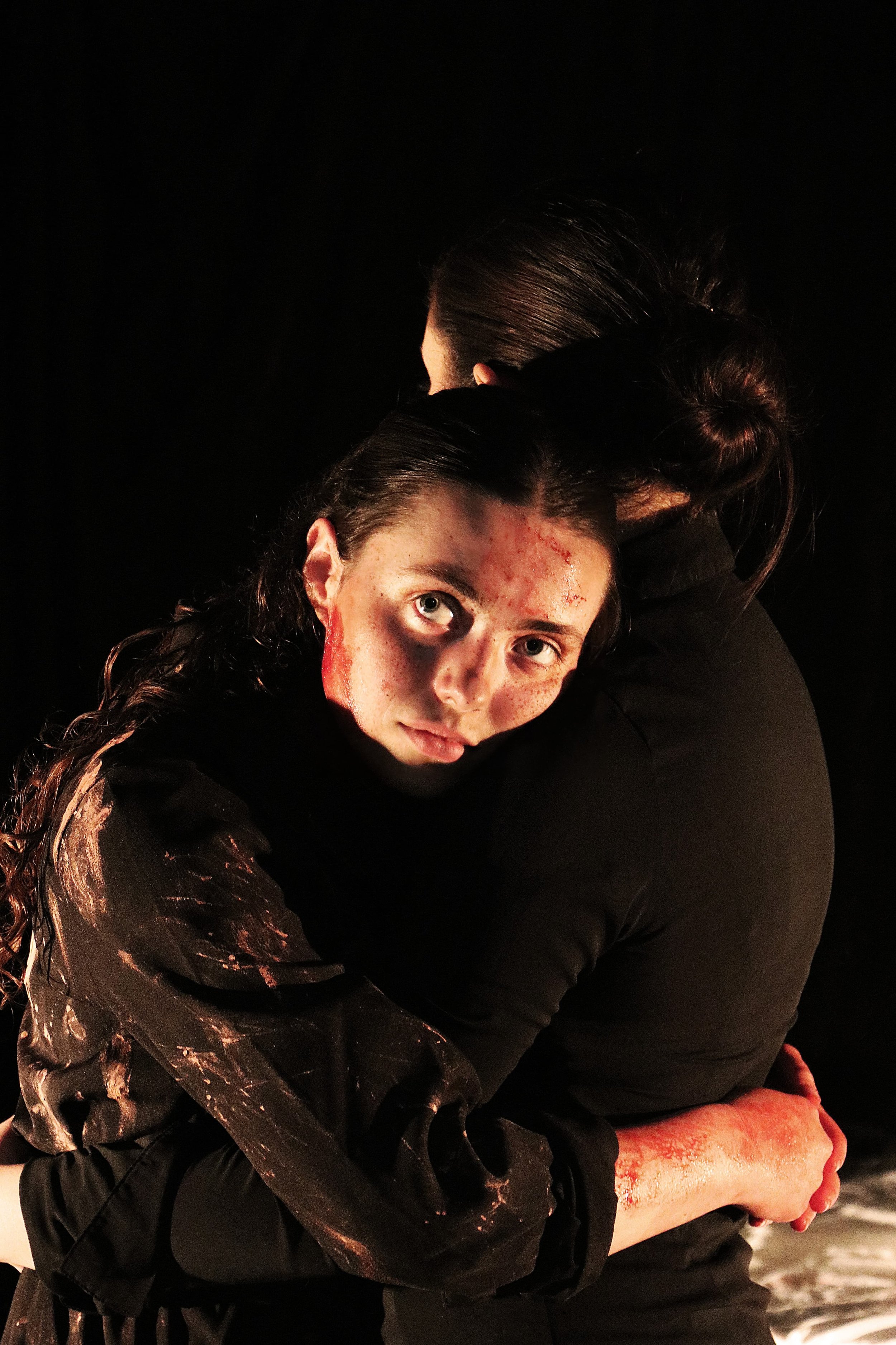 Two performers embrace in a dramatic stage scene, one with blood-like markings on her face and arm, both dressed in dark clothing against a black backdrop — Giles Forman Centre For Acting.