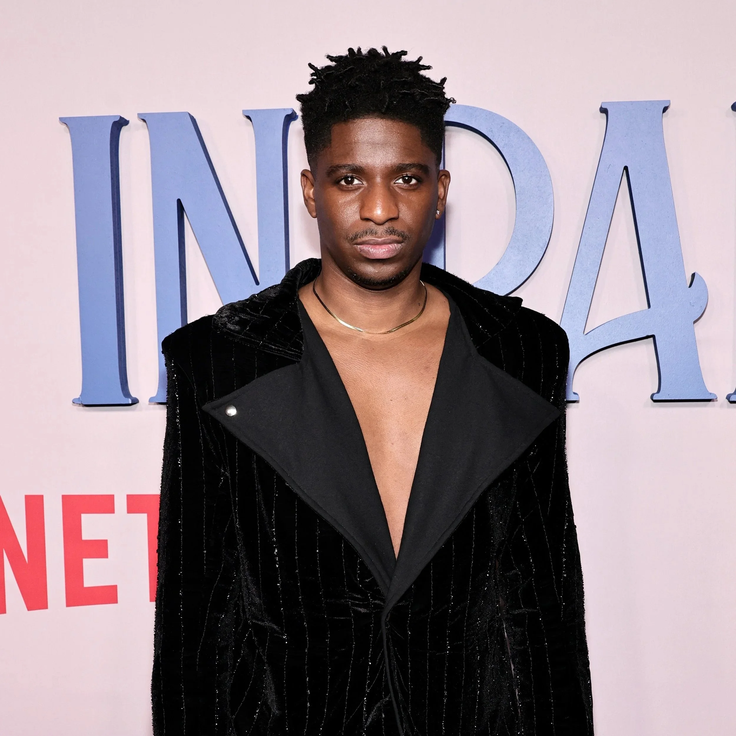 A young man with dark skin and short, curly hair stands in front of a light pink wall with large blue letters. He is wearing a black jacket, a black shirt, and a gold necklace, and looks directly at the camera.