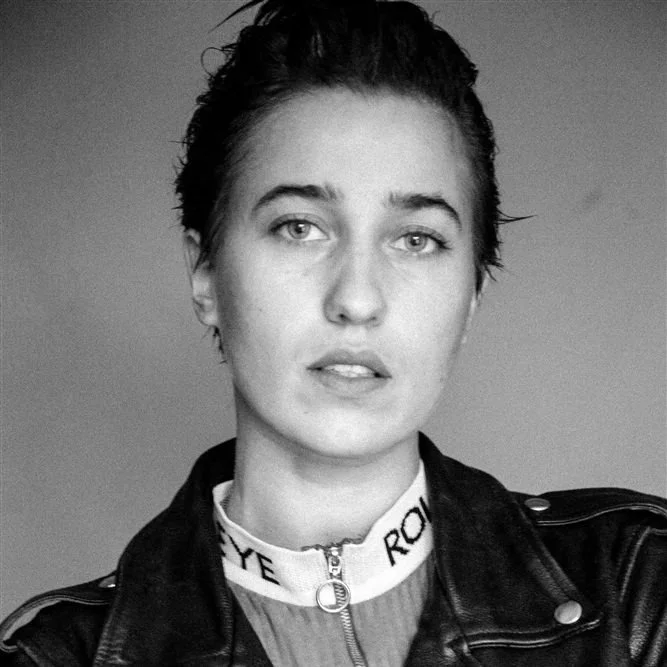 Black and white portrait of a young woman with short, wet hair, wearing a leather jacket and a high-collared shirt or top underneath.
