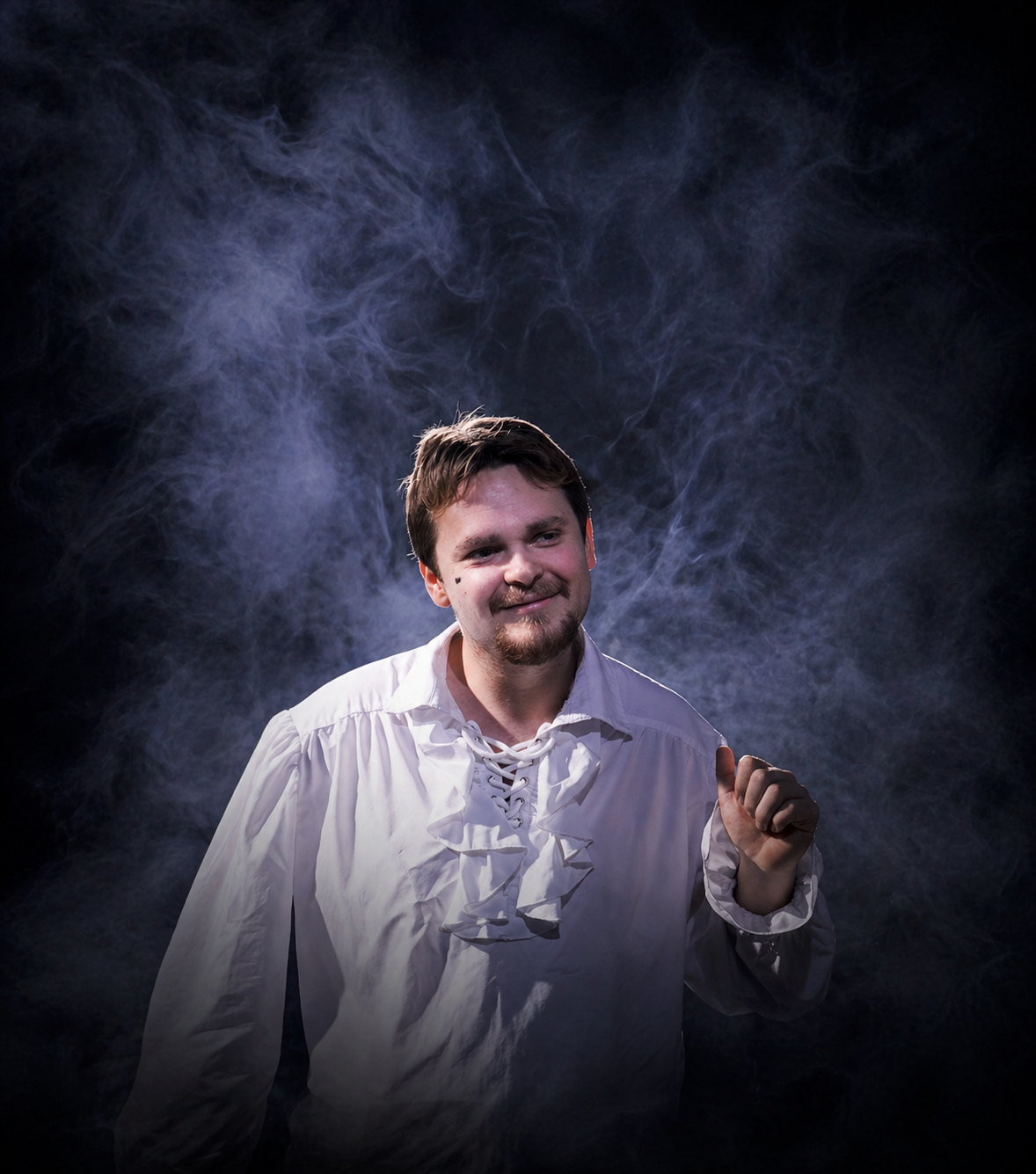 A man in a white shirt with ruffled front, smiling and looking to the side, surrounded by a smoky background.