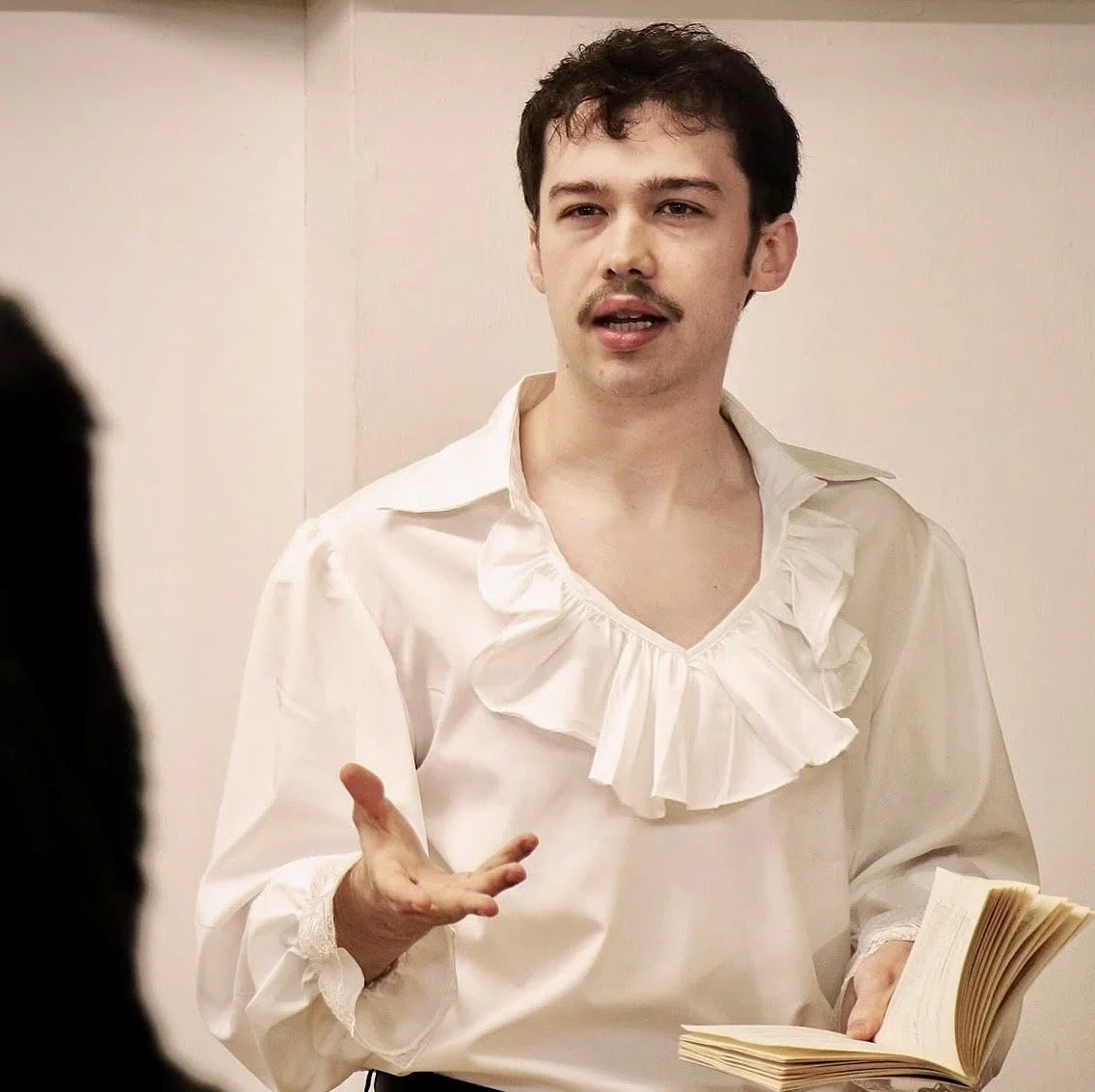 A man is talking with a slightly amused expression, holding an open book in his left hand, while gesturing with his right hand.
