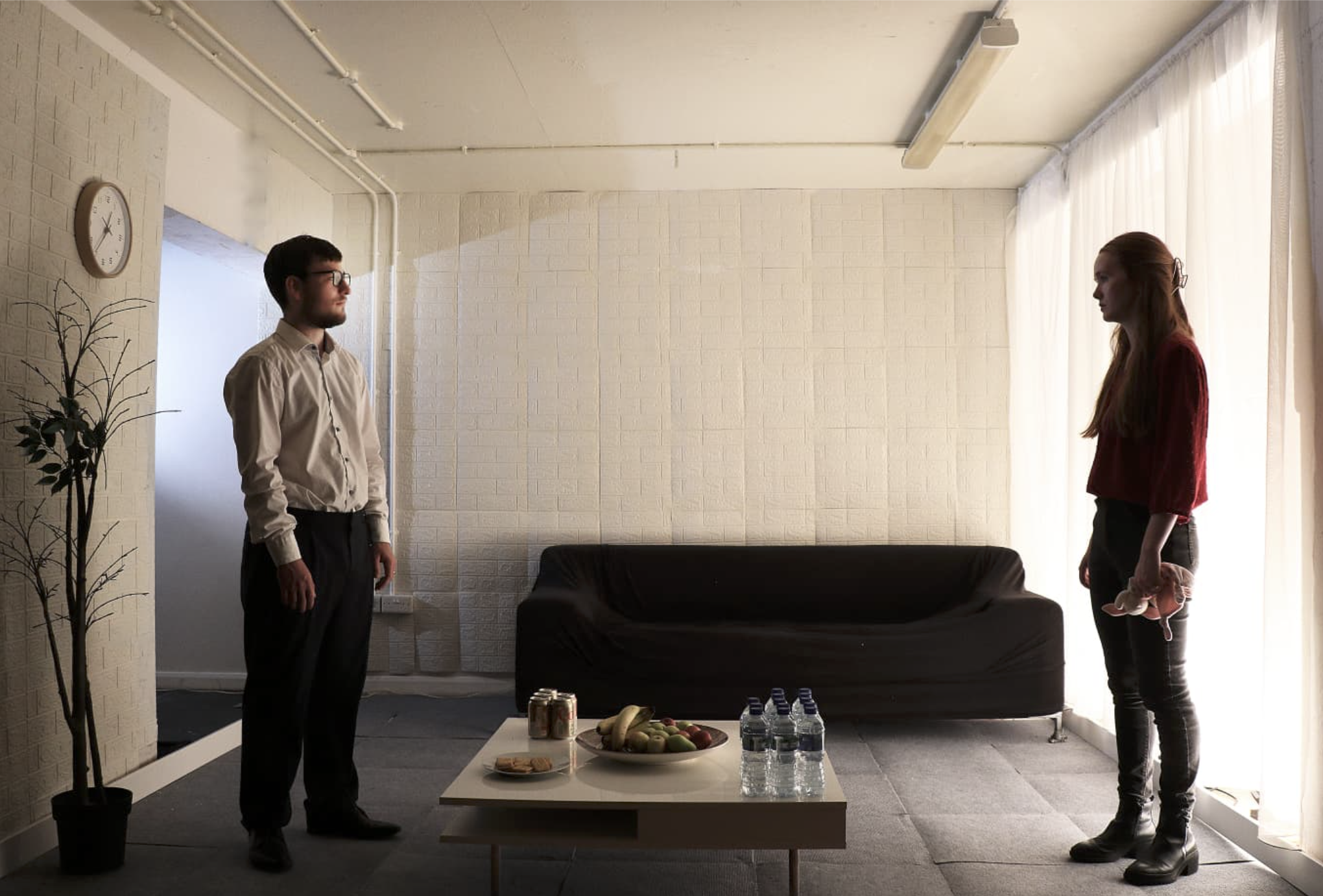 Two people standing in a room, facing each other. One man wearing glasses and a white shirt, and one woman with long red hair in a red top. There is a black couch, a table with water bottles, fruit, and other snacks, a wall clock showing 3:10, some p