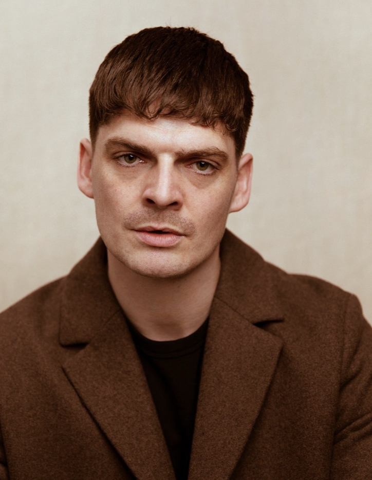 Portrait of a man with short brown hair wearing a brown blazer and black shirt, looking directly at the camera with a neutral expression.