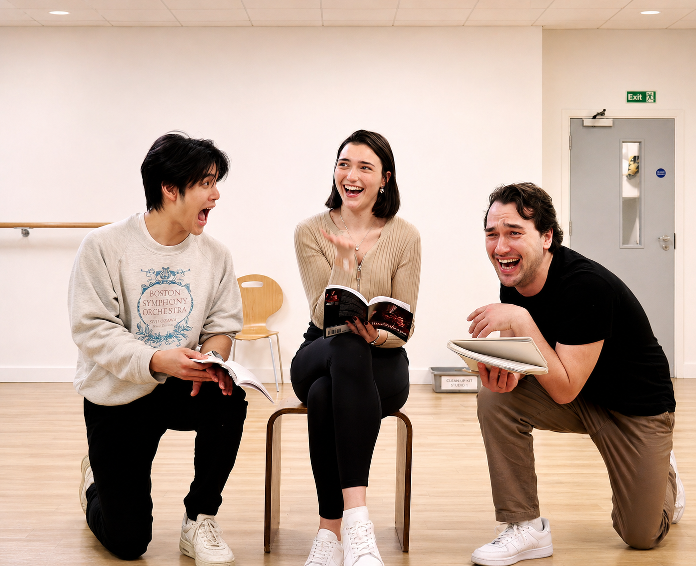 Three people engaging in a lively discussion in a dance studio, with two kneeling and one sitting on a chair, all smiling and holding books or scripts.