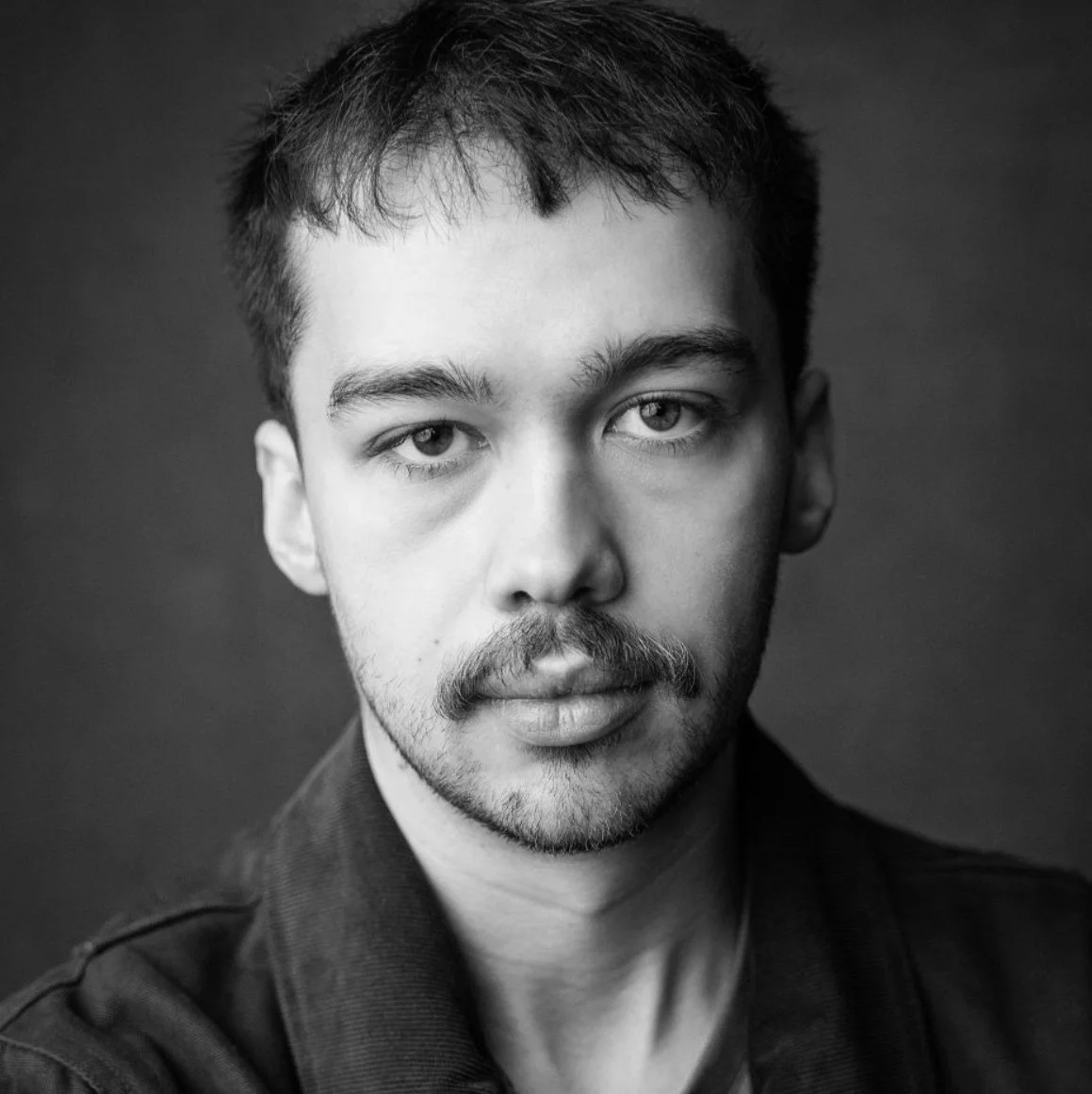 A black and white close-up portrait of a young man with dark hair, light eyes, facial hair, and wearing a dark shirt, looking directly at the camera.