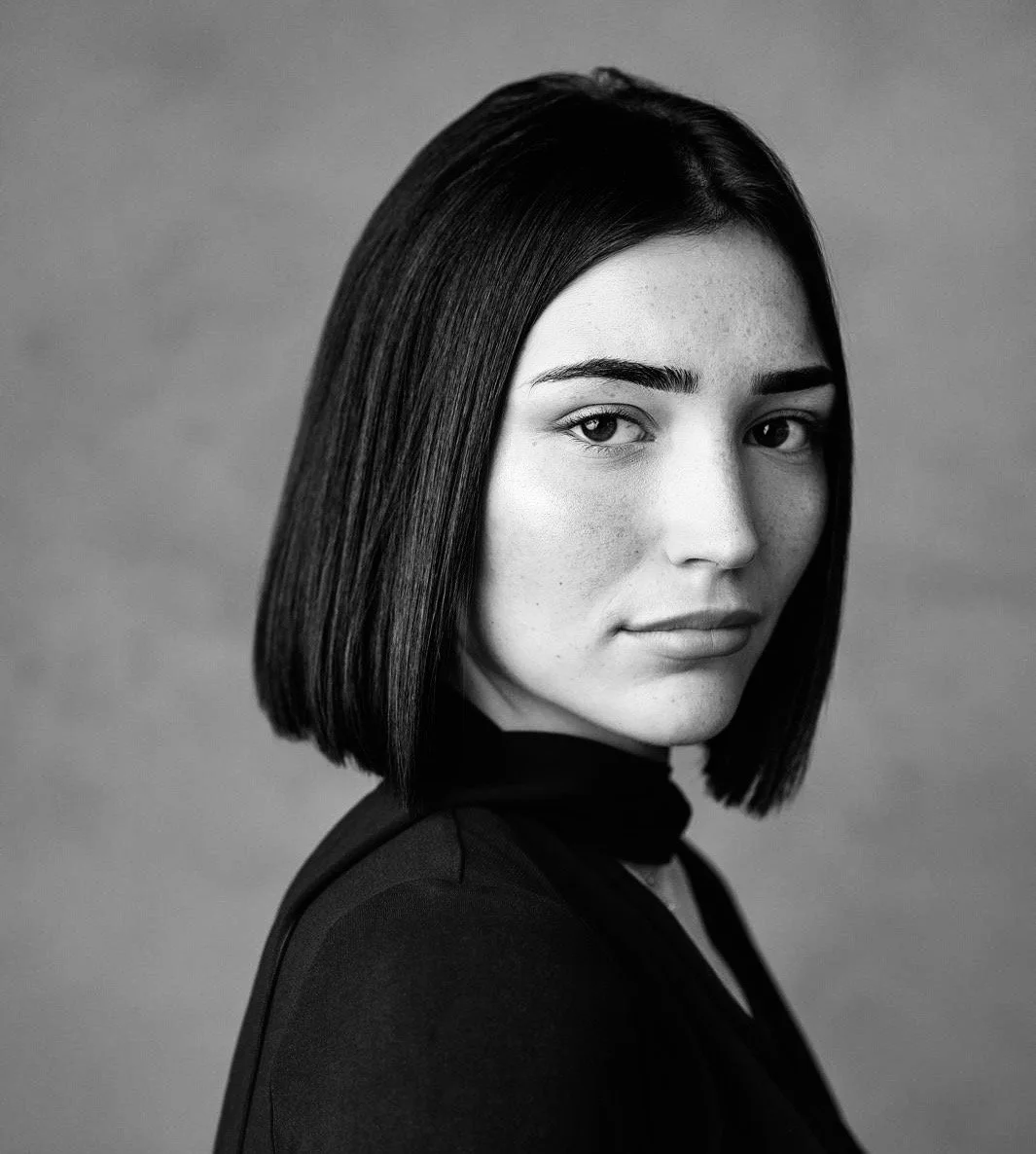 Black and white portrait of a woman with straight, shoulder-length dark hair, wearing a dark top with a cut-out neckline, looking at the camera with a neutral expression.