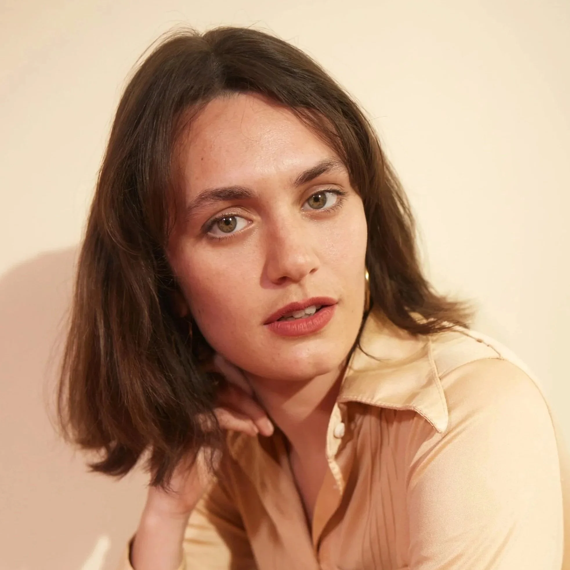 A woman with brown hair and green eyes wearing a beige or light-colored shirt or dress, posing with her hand resting on the side of her neck against a plain light background.