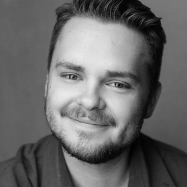 Black and white portrait of a young man with short hair, a beard, and a friendly smile, looking directly at the camera.