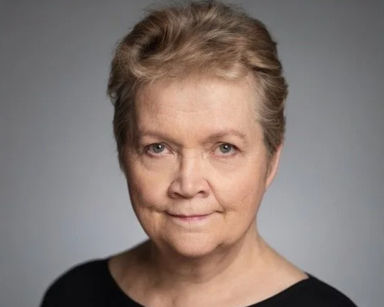 Close-up portrait of an older woman with short, light brown hair and blue eyes, wearing a black top against a plain gray background.