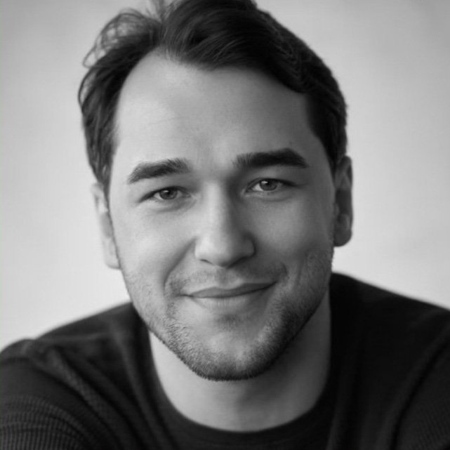 Black and white portrait of a smiling young man with short dark hair, light eyes, and light facial hair, wearing a dark shirt, against a plain light background.