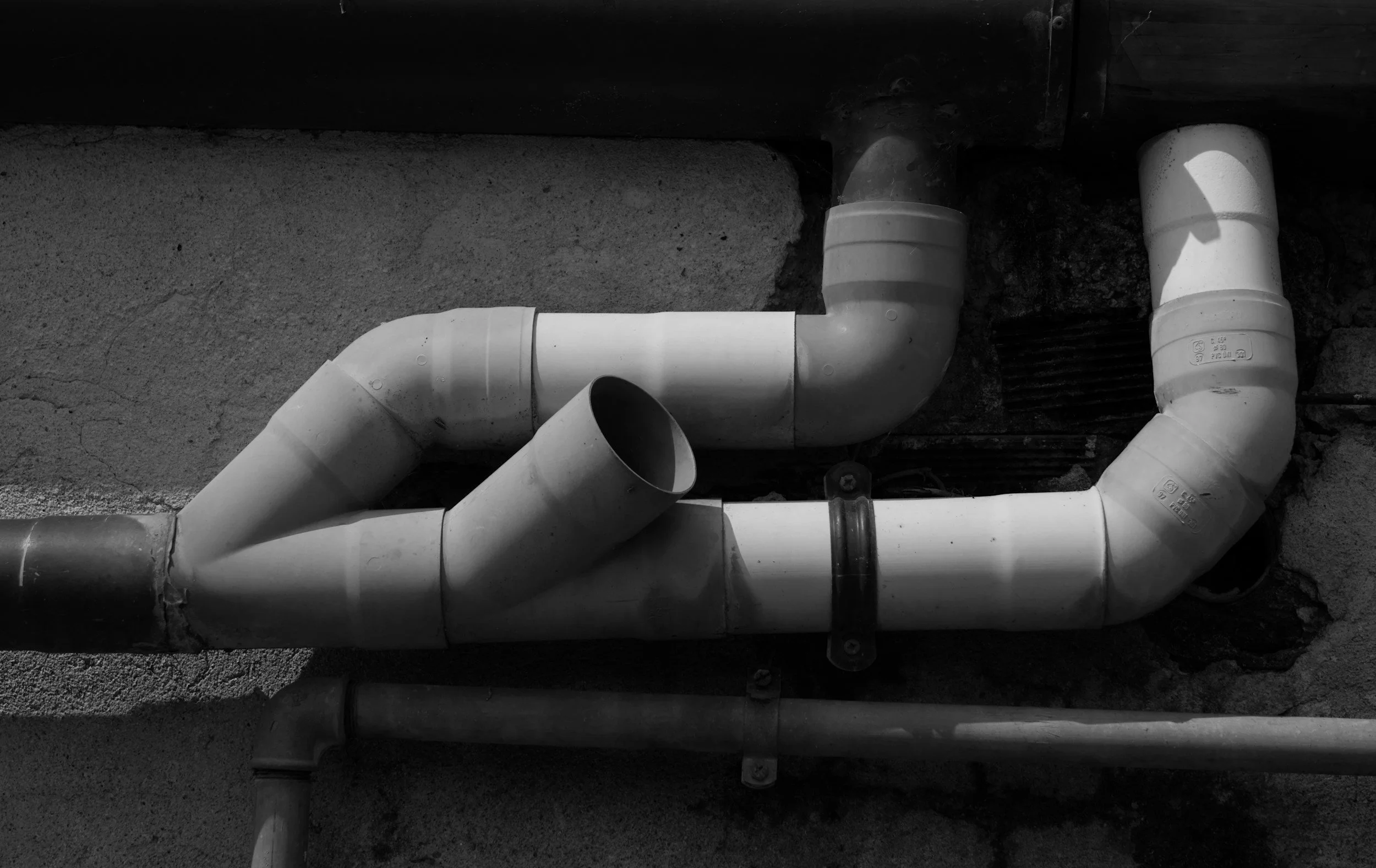 Black and white photo of plumbing pipes on an exterior wall, consisting of several white and black PVC pipes connected with elbows and fittings.