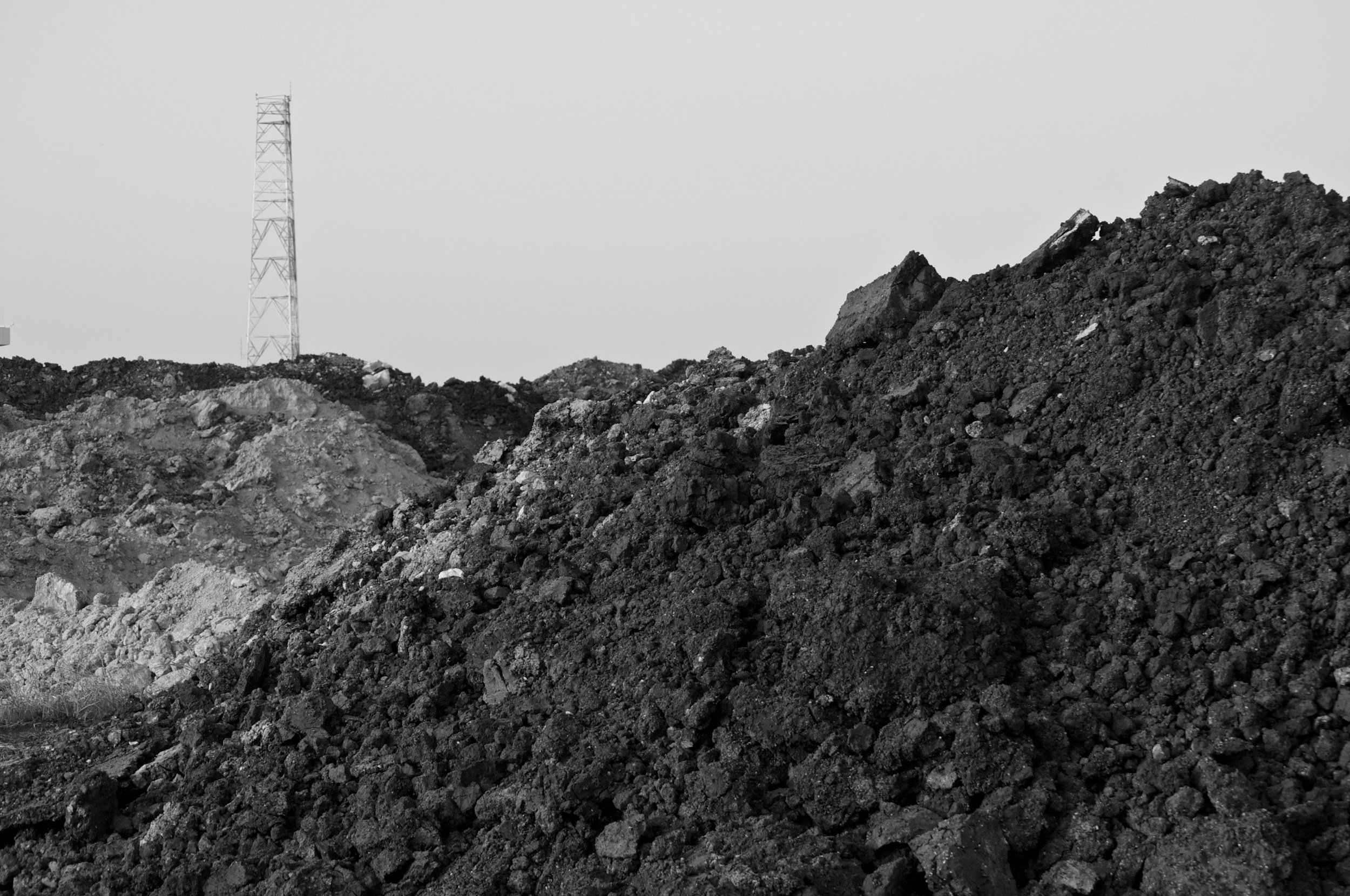 Pile of dark dirt or coal in the foreground with an electrical tower in the background under a clear sky.