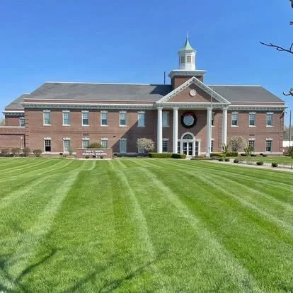 A large brick building with a clock and a steeple, surrounded by a well-maintained green lawn and a clear blue sky.
