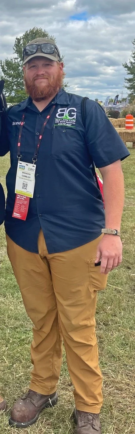 Man wearing a navy blue shirt with logos, khaki pants, and boots, standing outdoors with trees and cloudy sky in the background, at an event with banners and hay bales.
