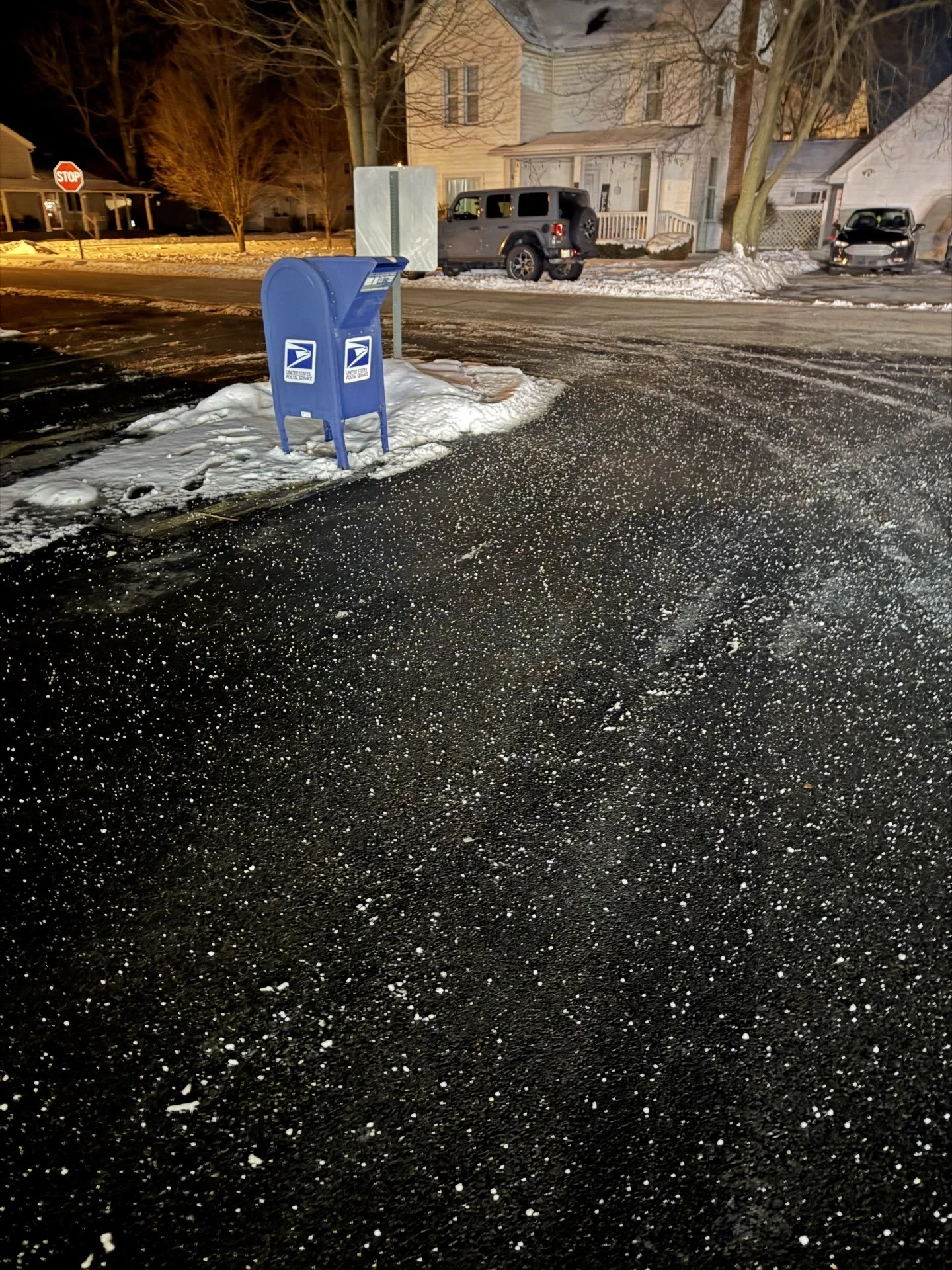 Nighttime scene of a residential neighborhood with a street, parked cars, snow on the ground, and a blue USPS mailbox in the foreground.
