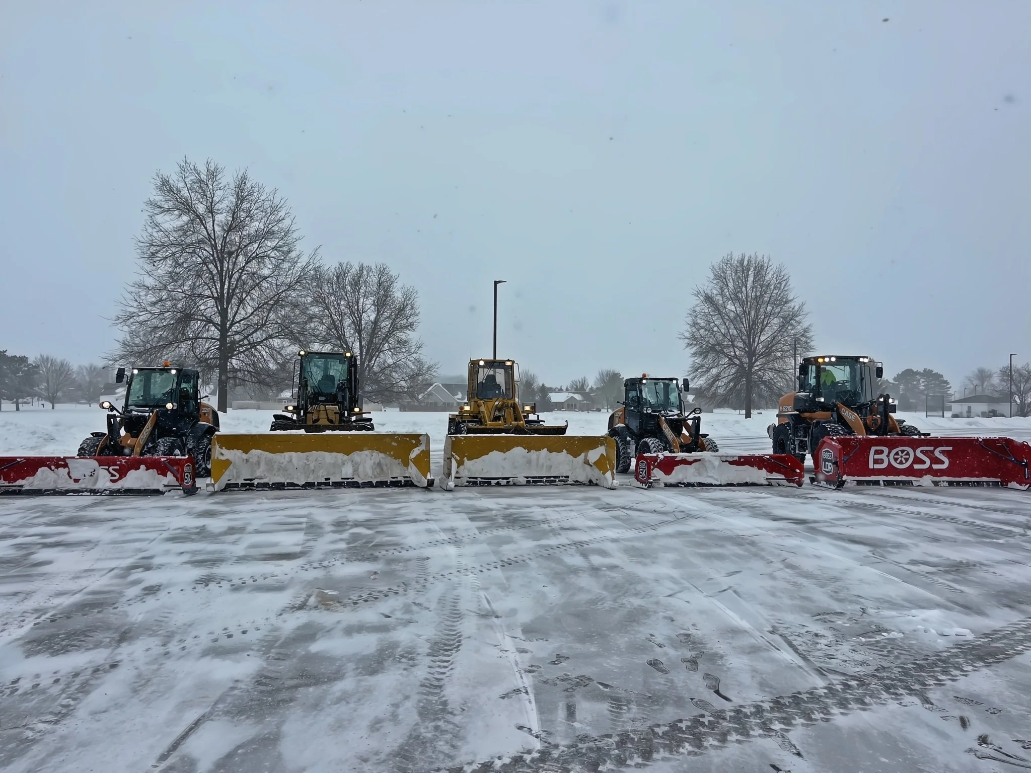 Five snow plow trucks lined up on a snow-covered parking lot with trees and houses in the background, snowy sky overhead.