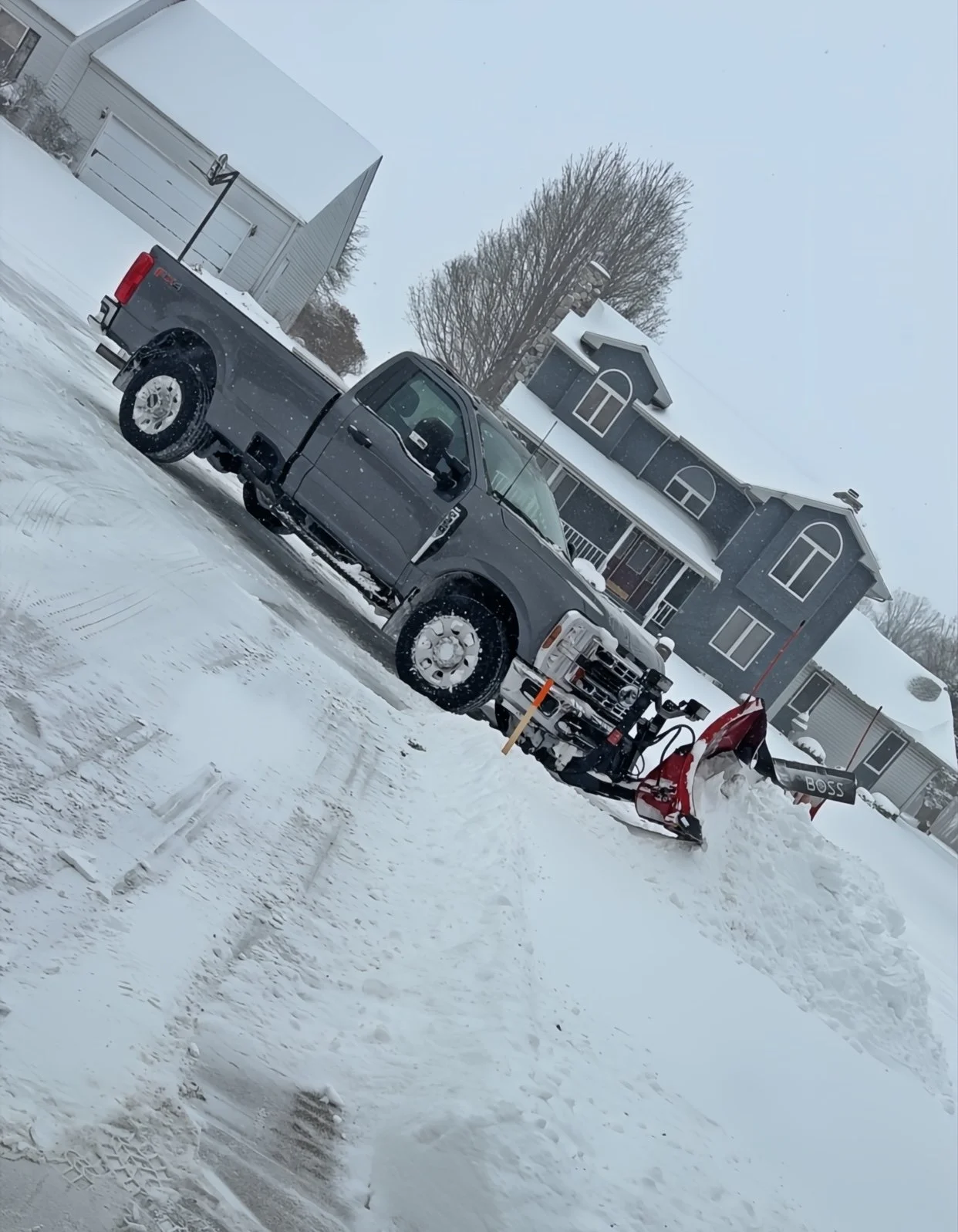 A black pickup truck with a snow plow attached to the front clearing snow on a residential street during winter weather.