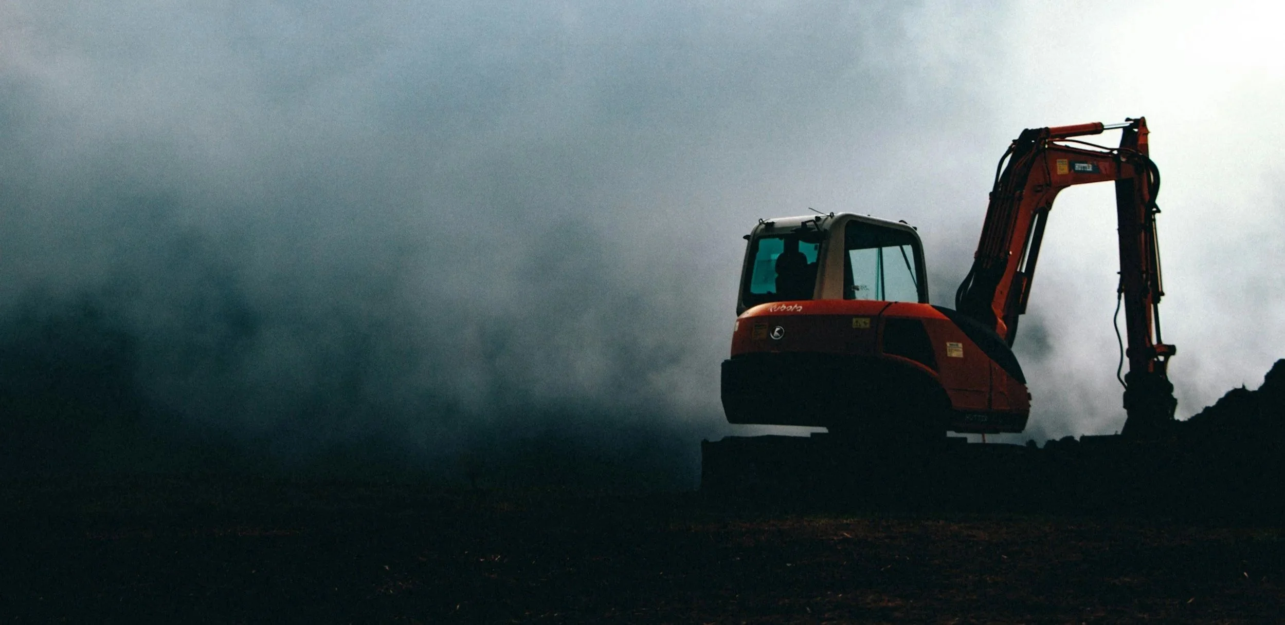 A silhouette of an orange excavator against a dark, cloudy sky on a construction site.