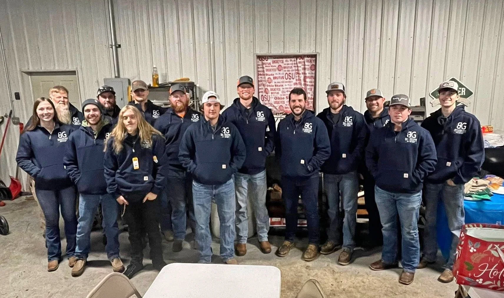 Group of 14 people standing indoors in front of a wall, all wearing matching dark blue jackets with a logo, smiling at the camera.