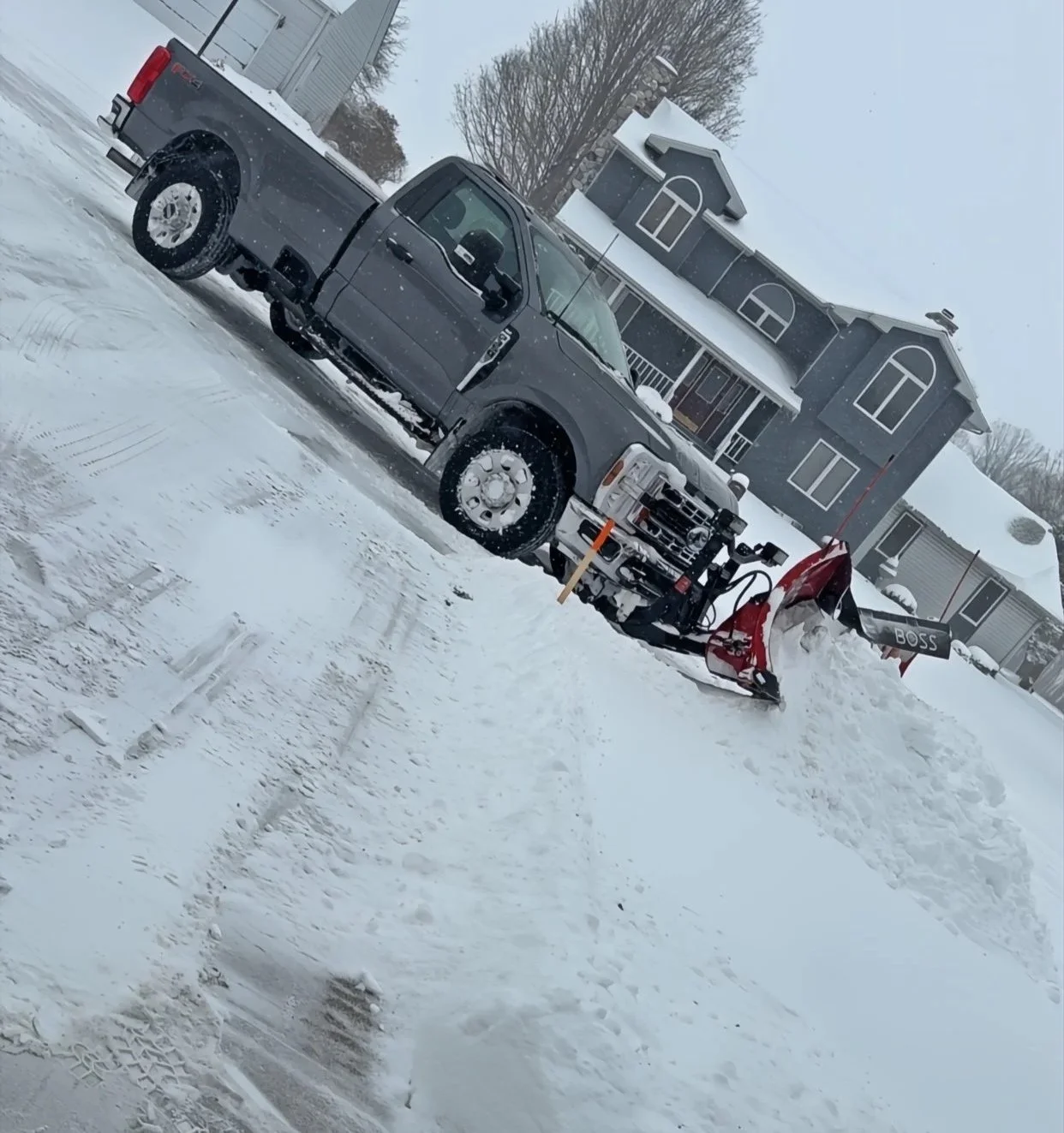 A gray pickup truck equipped with a snow plow blade attached to the front, parked on a snow-covered driveway in front of a black house with white trim in a snowy setting.