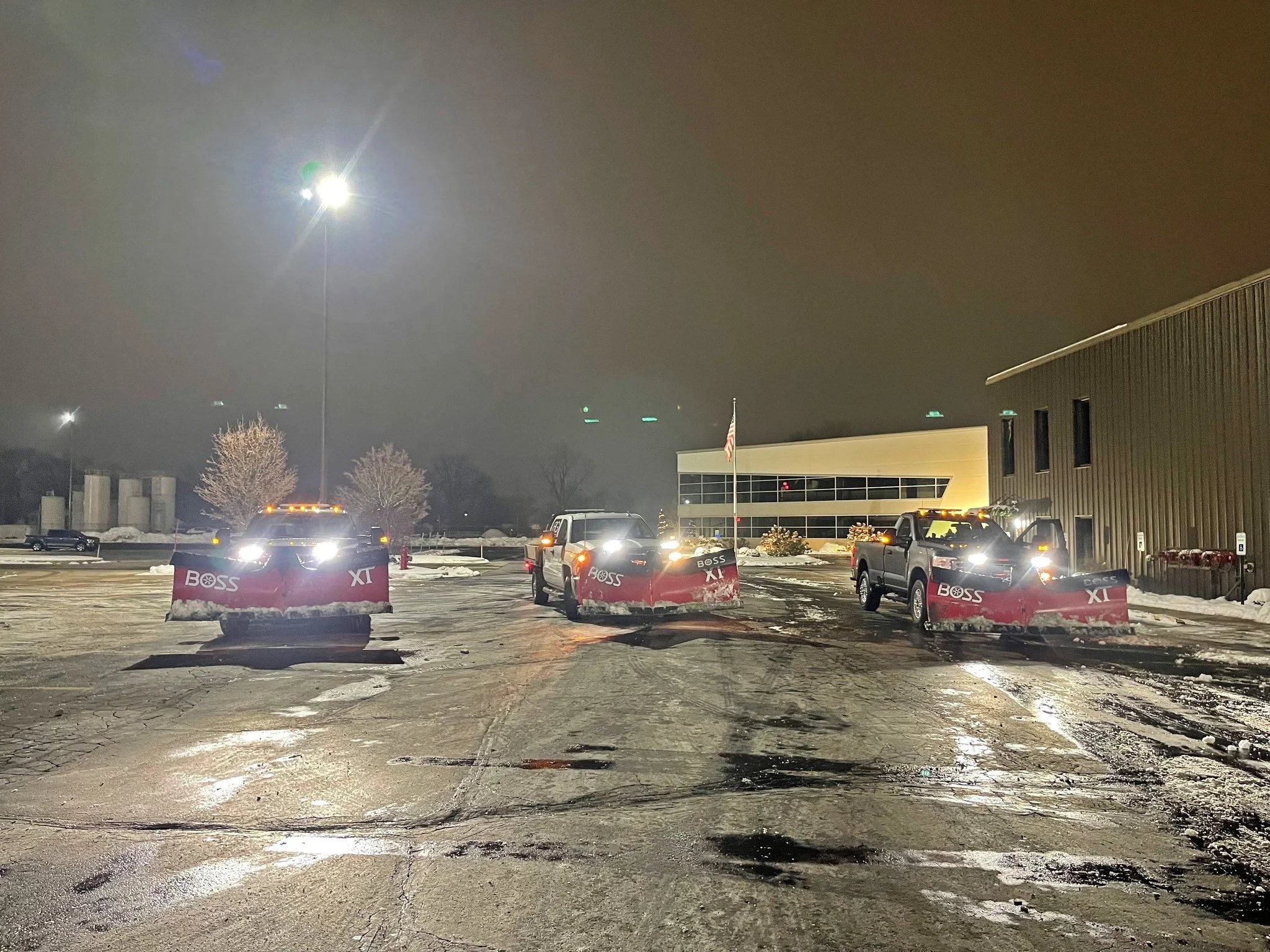 Three trucks equipped with snow plows parked in a snow-covered parking lot at night, illuminated by streetlights, with a building and an American flag in the background.