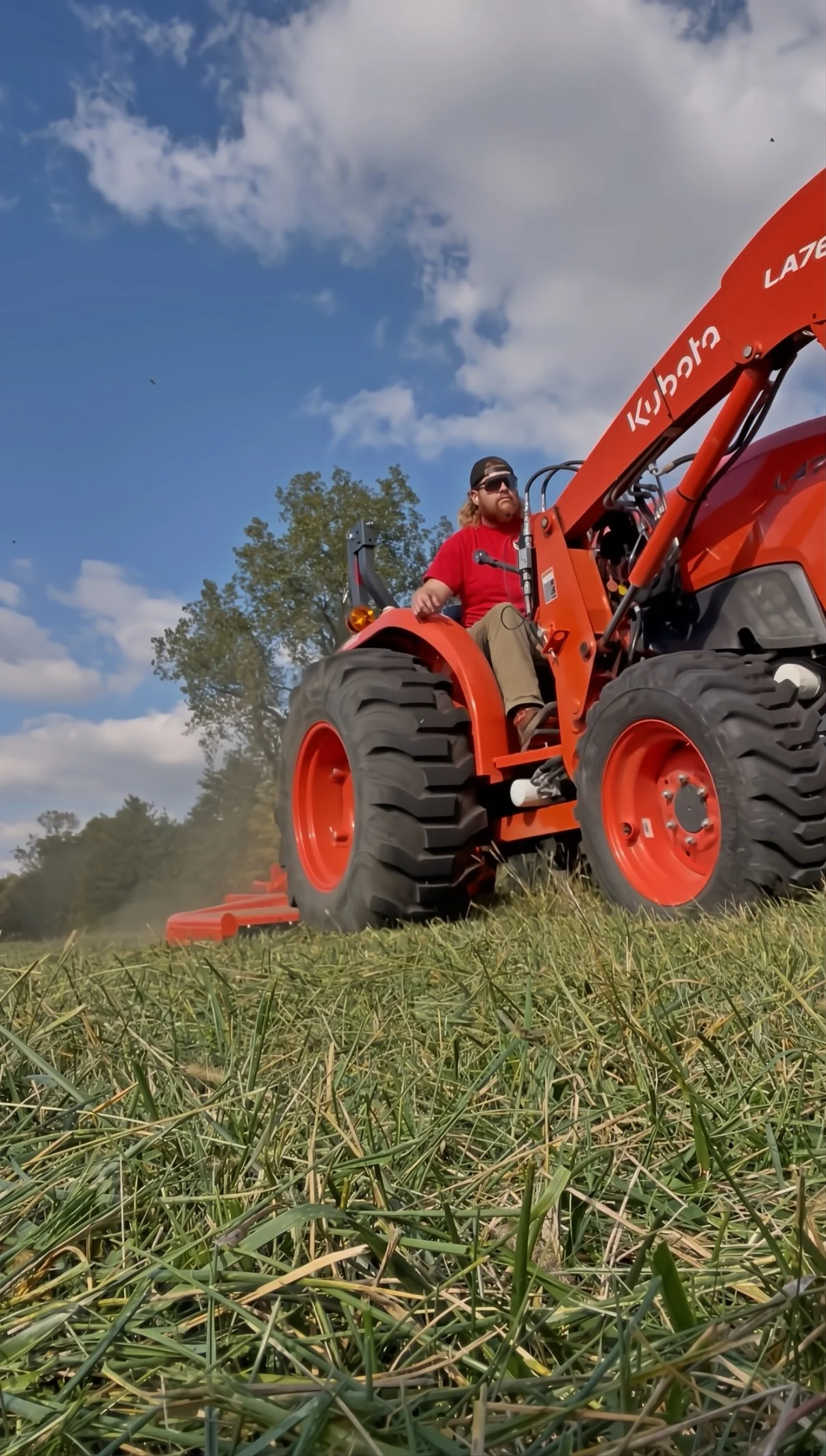 Man operating a red tractor on a grassy field on a sunny day with some clouds in the sky.