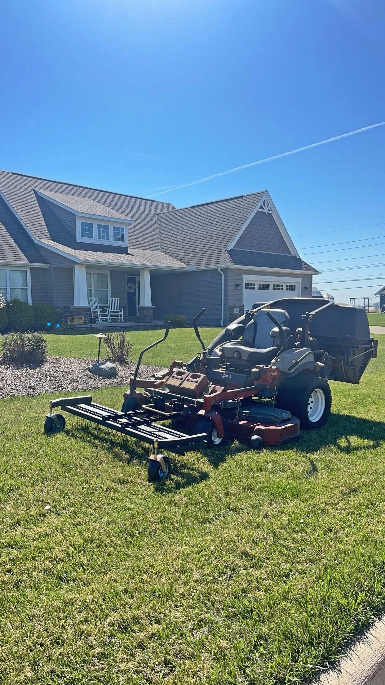 A riding lawn mower on a grassy lawn in front of a gray house with a porch, under a clear blue sky.