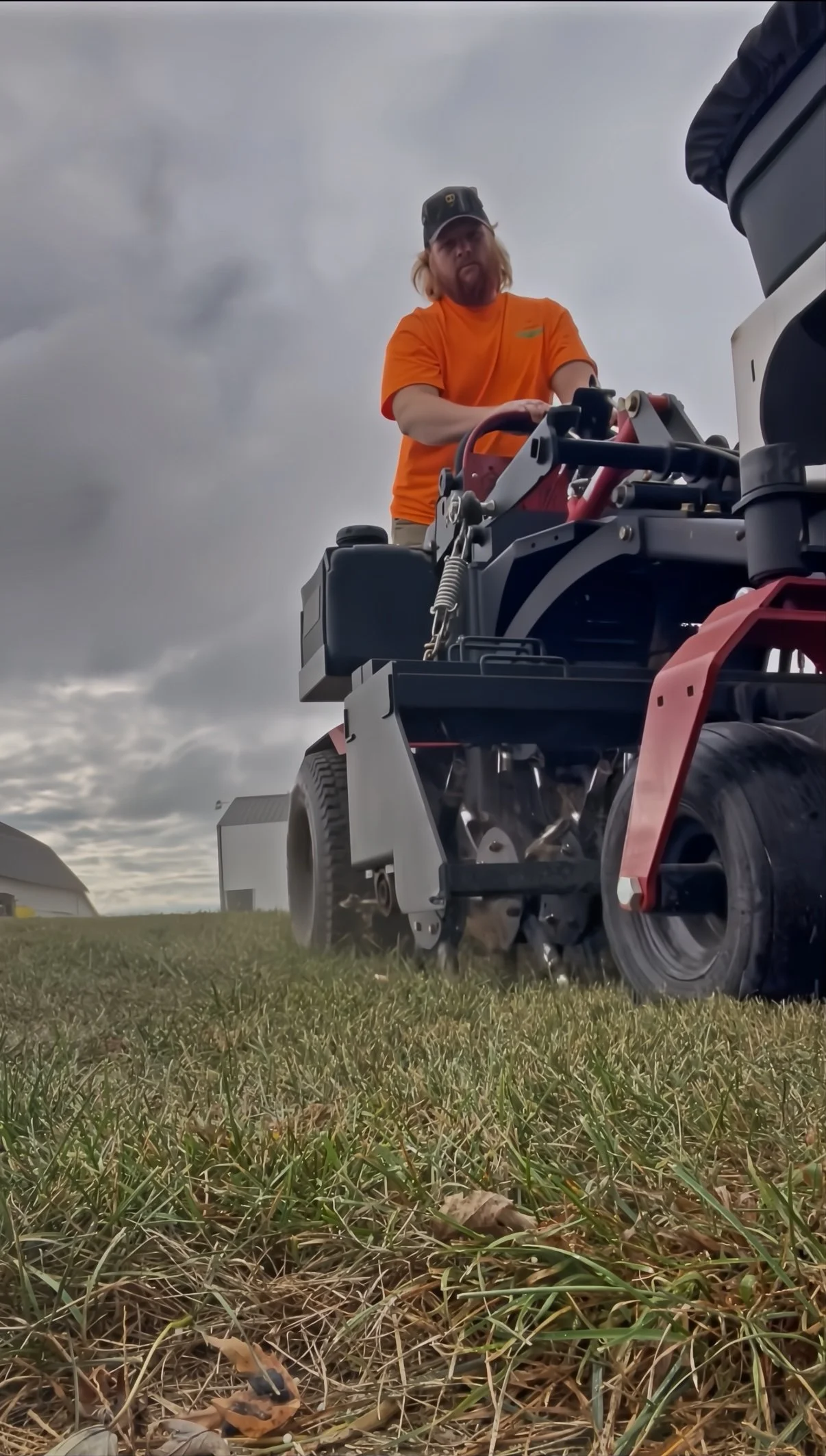 A person with long blond hair, wearing an orange shirt and a dark cap, operating a drive-on lawn mower on a grassy field with a cloudy sky in the background.