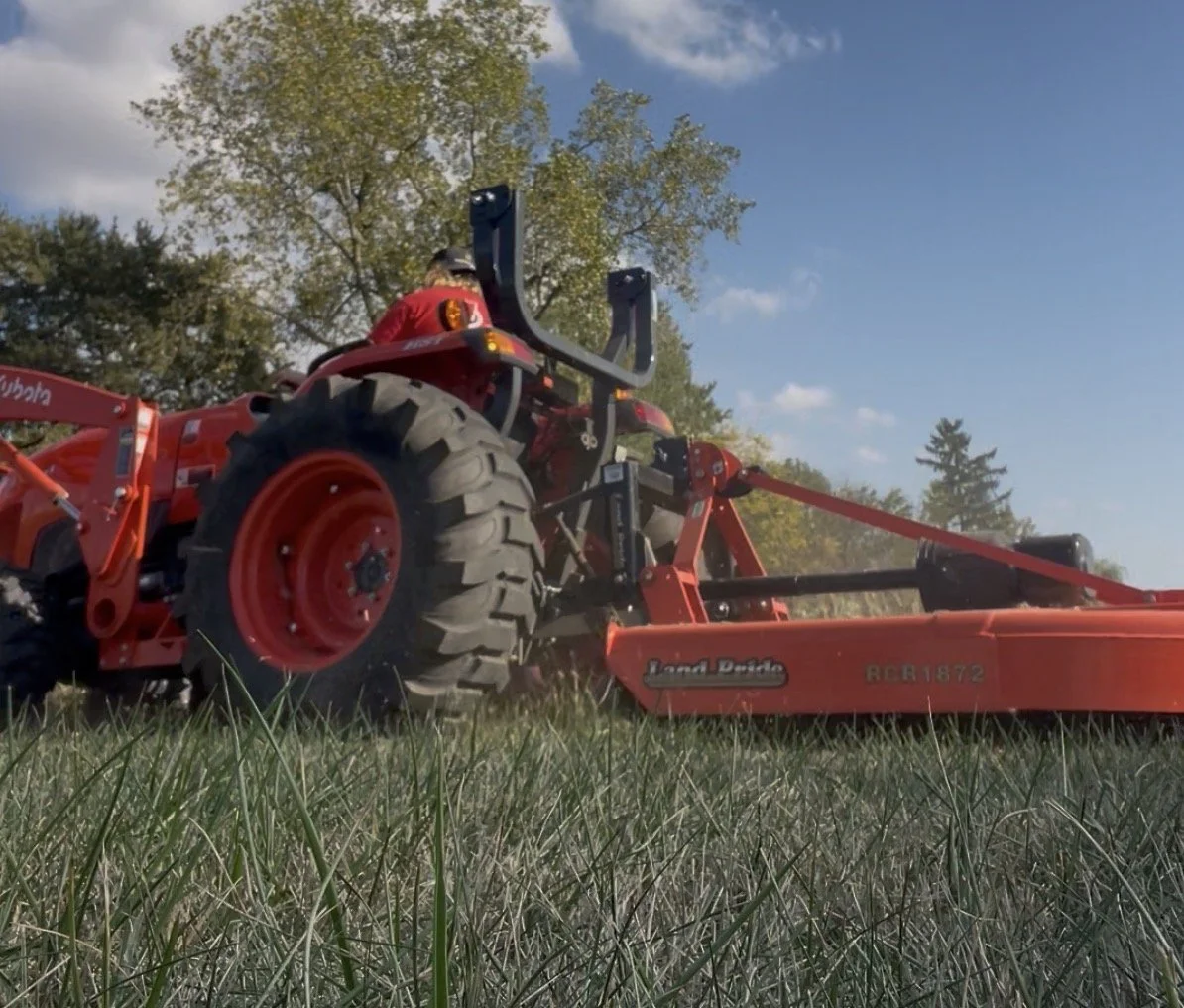 A person operating a red tractor in a grassy field with trees in the background under a blue sky with some clouds.
