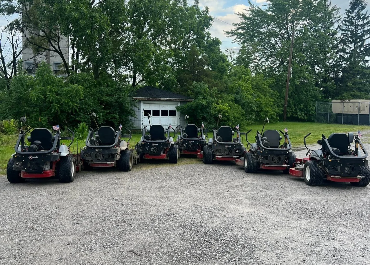 Six riding lawn mowers parked in a row on a gravel lot with green trees and a small white shed in the background.