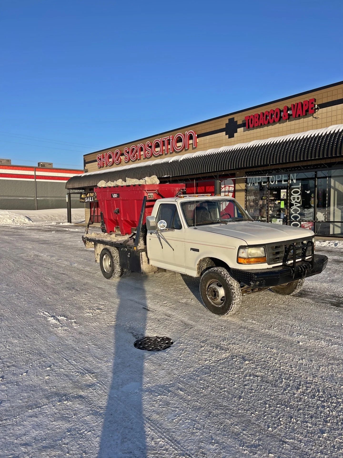 A white utility truck with a red container loaded with snow, parked outside a shoe store named 'Shoe Sensation' that sells tobacco and vape products, in a snowy parking lot with a clear blue sky.