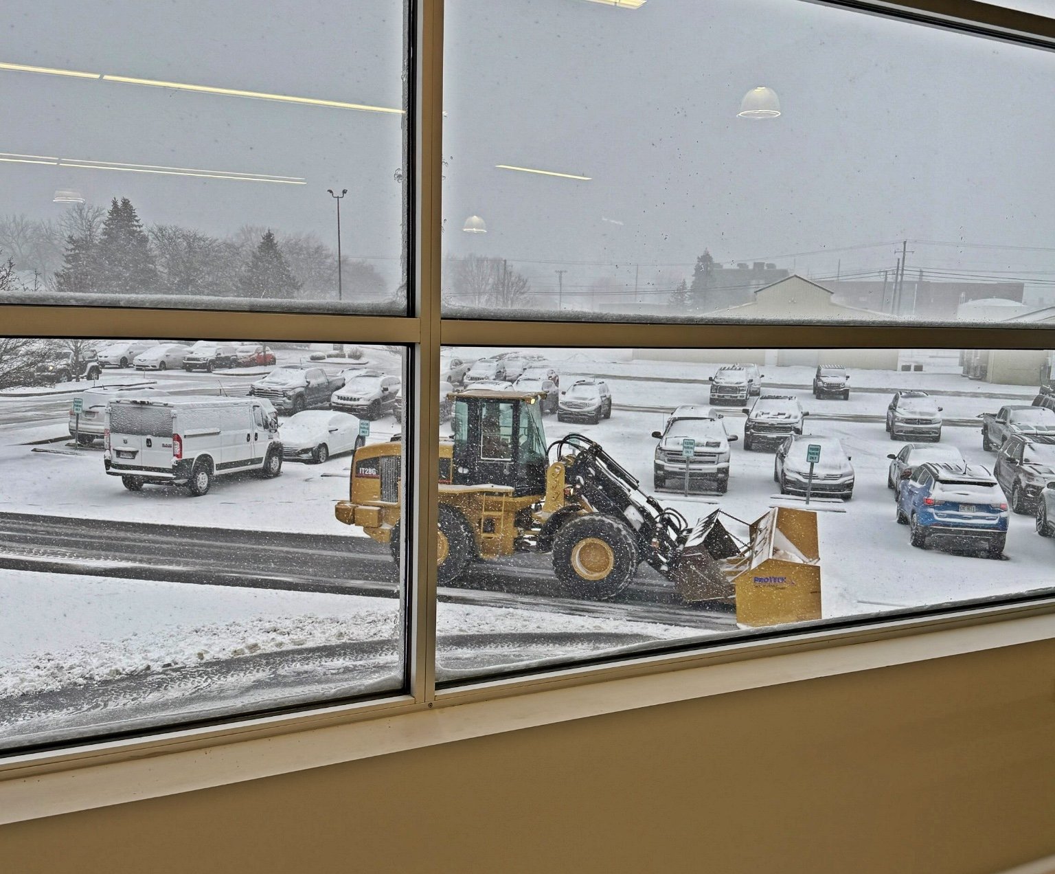 View through a window shows a snow-covered parking lot with a yellow snow plow clearing the snow, surrounded by parked cars and vehicles during snowy weather.