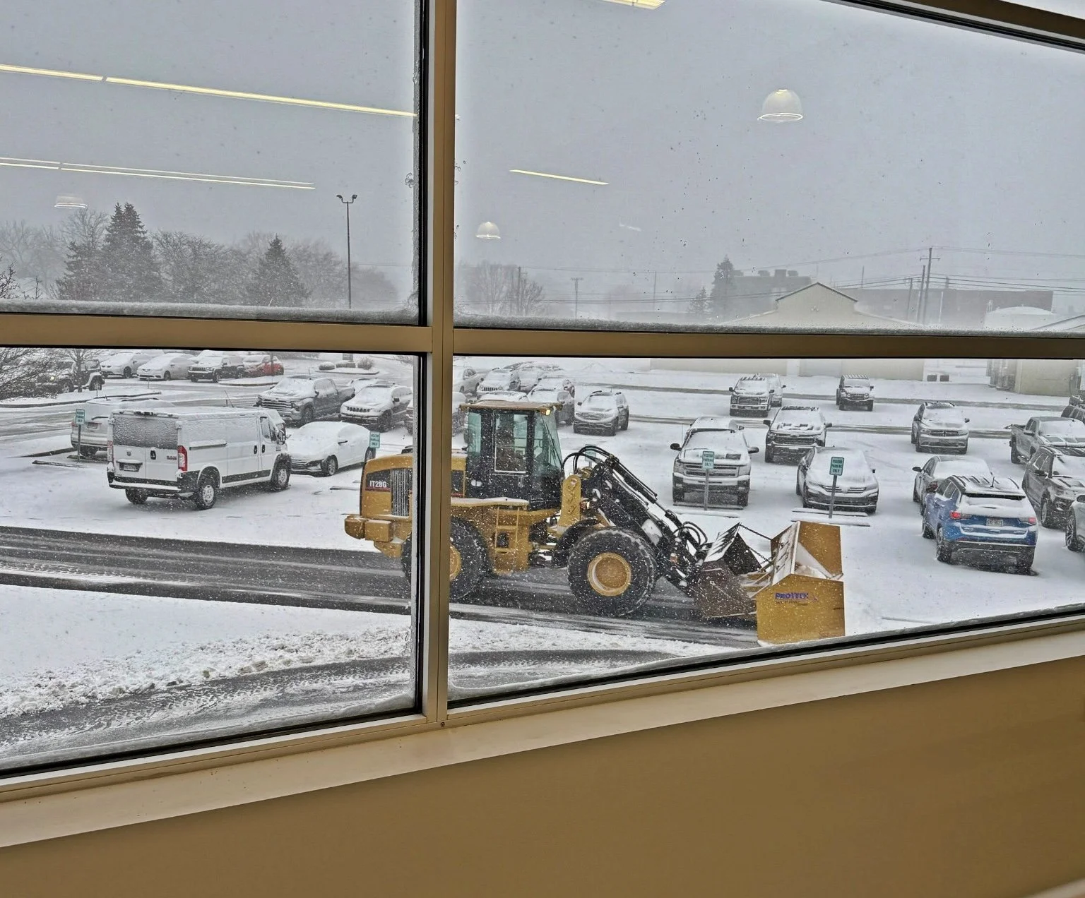 Snow removal vehicle clearing snow from a parking lot, seen through window panes with snow-covered cars and parking meters.