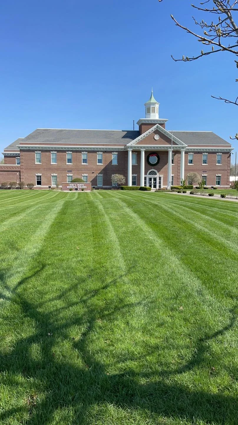 Large brick building with a clock tower and a wreath on the front, surrounded by a well-maintained lawn with small trees and bushes, clear blue sky overhead.