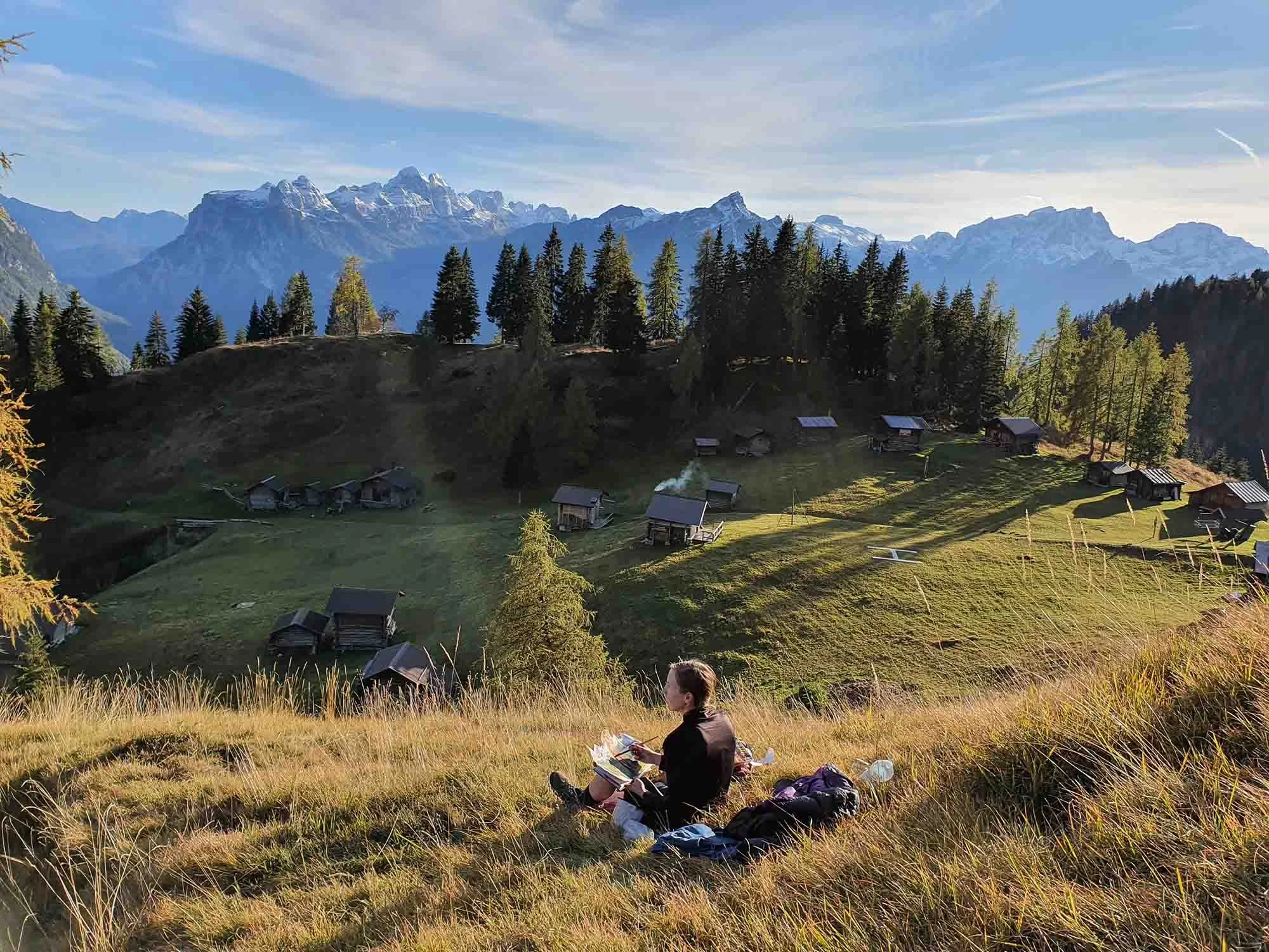 Ellie Middleton Illustrator painting in the mountains on location. She holds a paintbrush in one hand and has her sketchbook on her lap. Surrounded by the Italian Dolomites mountains